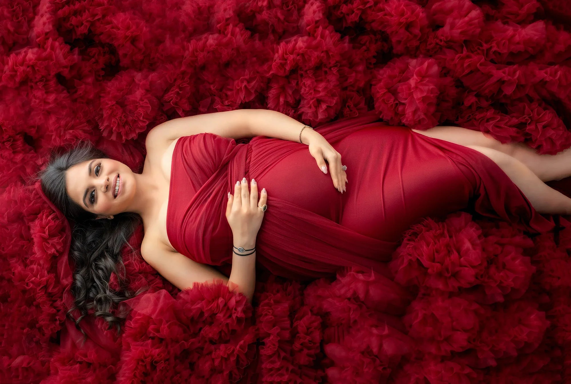 Pregnant woman in elegant red maternity gown lying on ruffled fabric, smiling