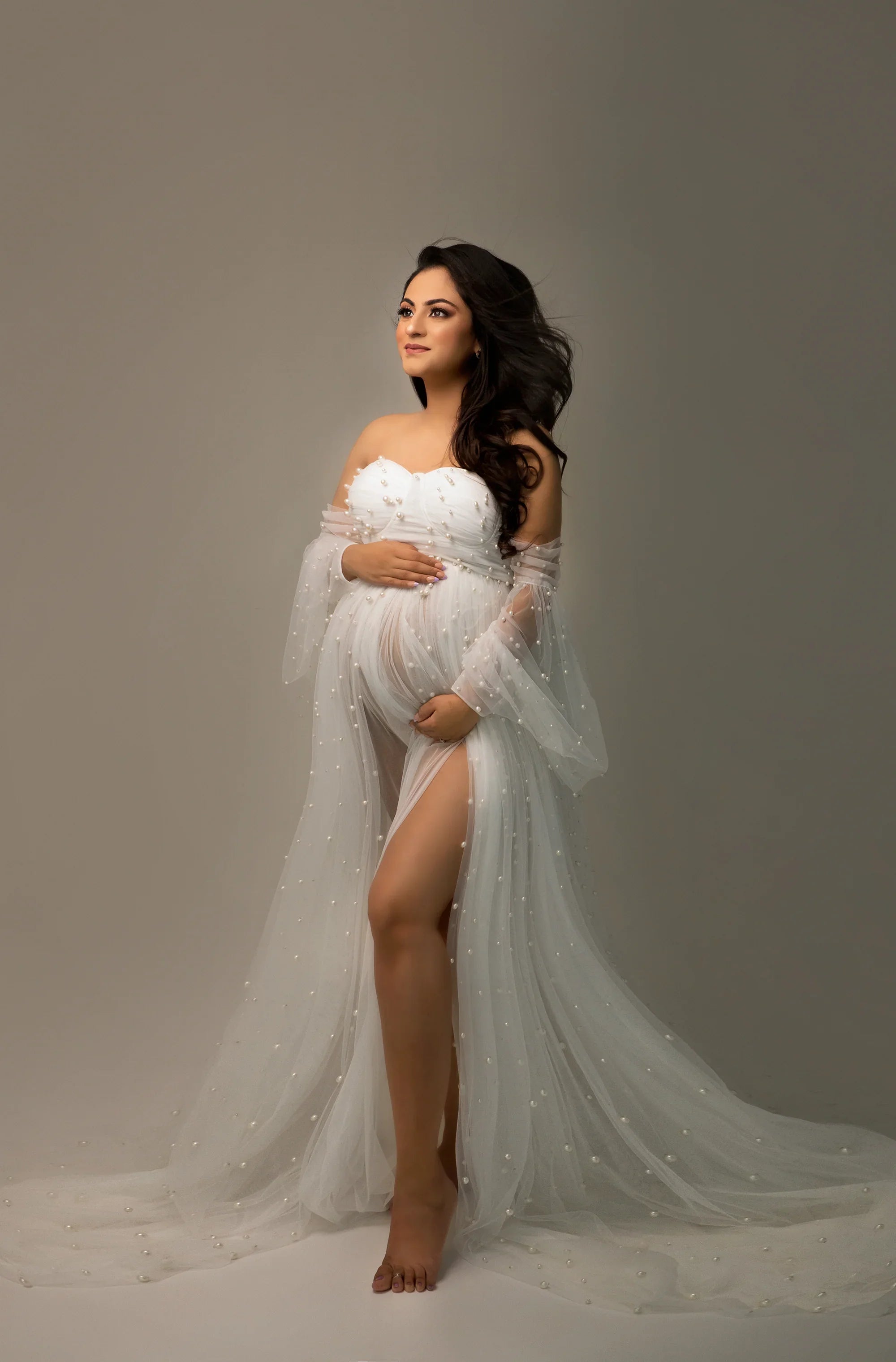 Pregnant woman in elegant white sheer maternity gown with pearl accents, holding belly