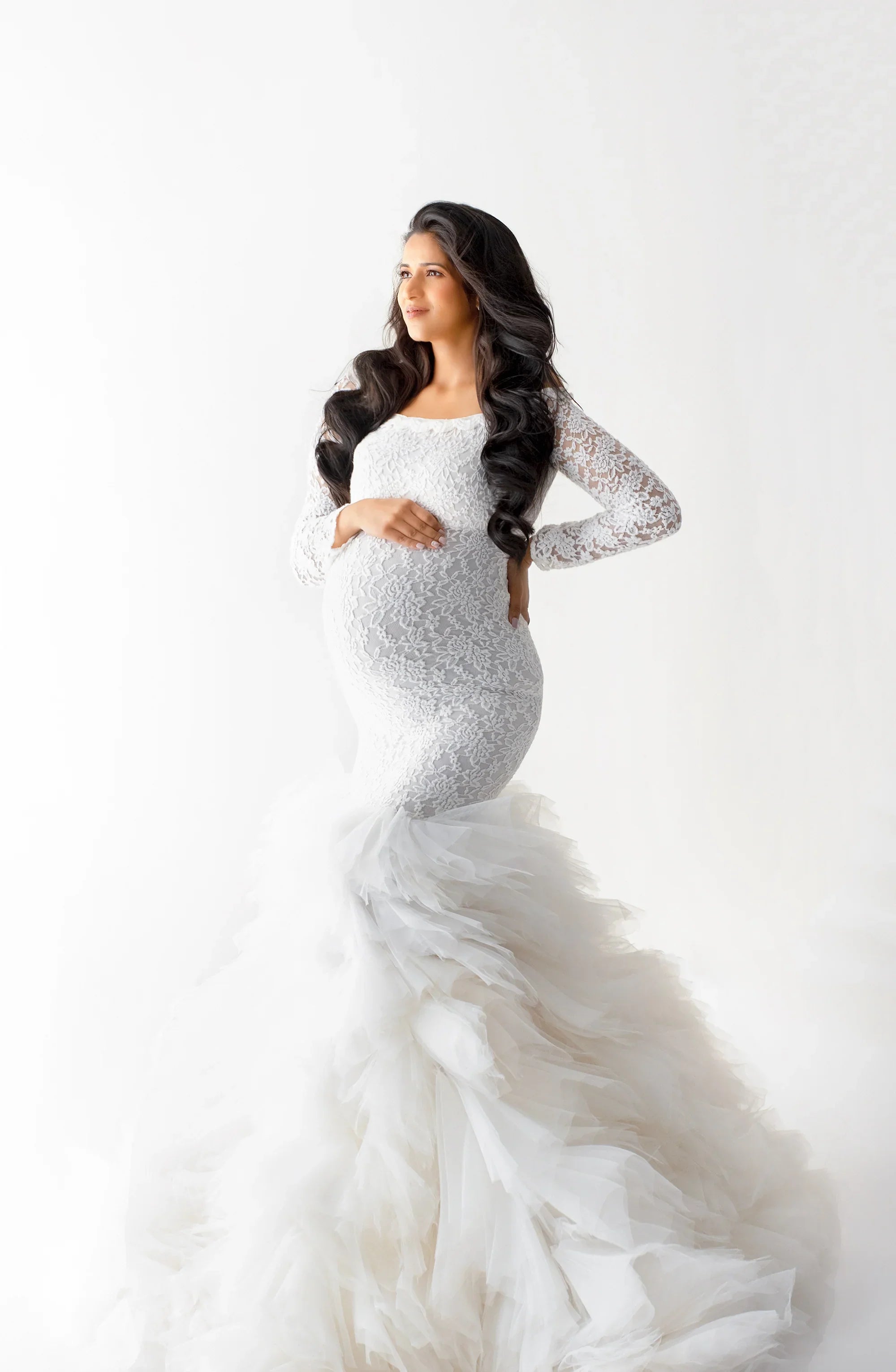 Pregnant woman in elegant white lace maternity gown with tulle train, posing on white backdrop