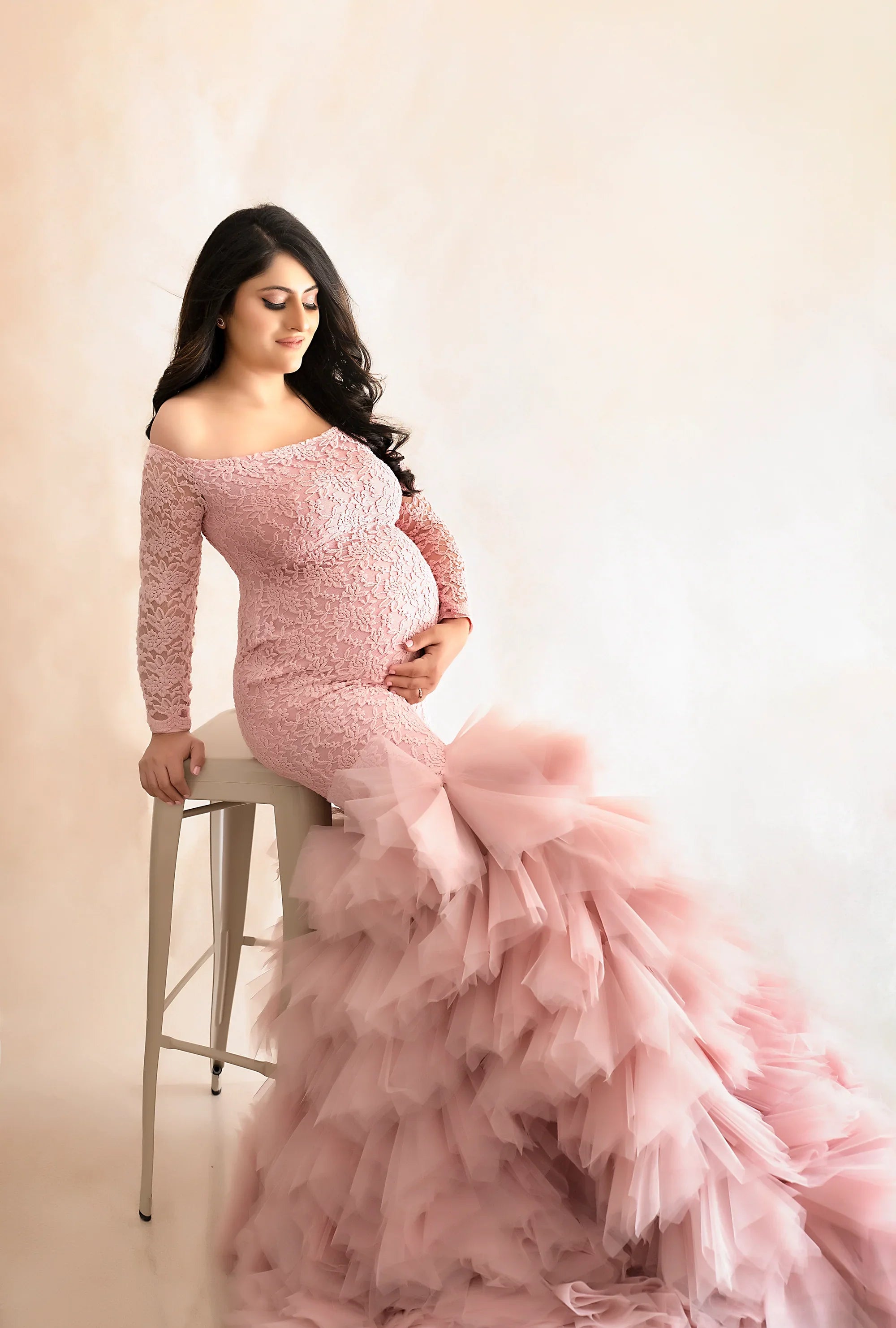 Pregnant woman in elegant pink lace maternity gown with tulle ruffles, sitting on a stool
