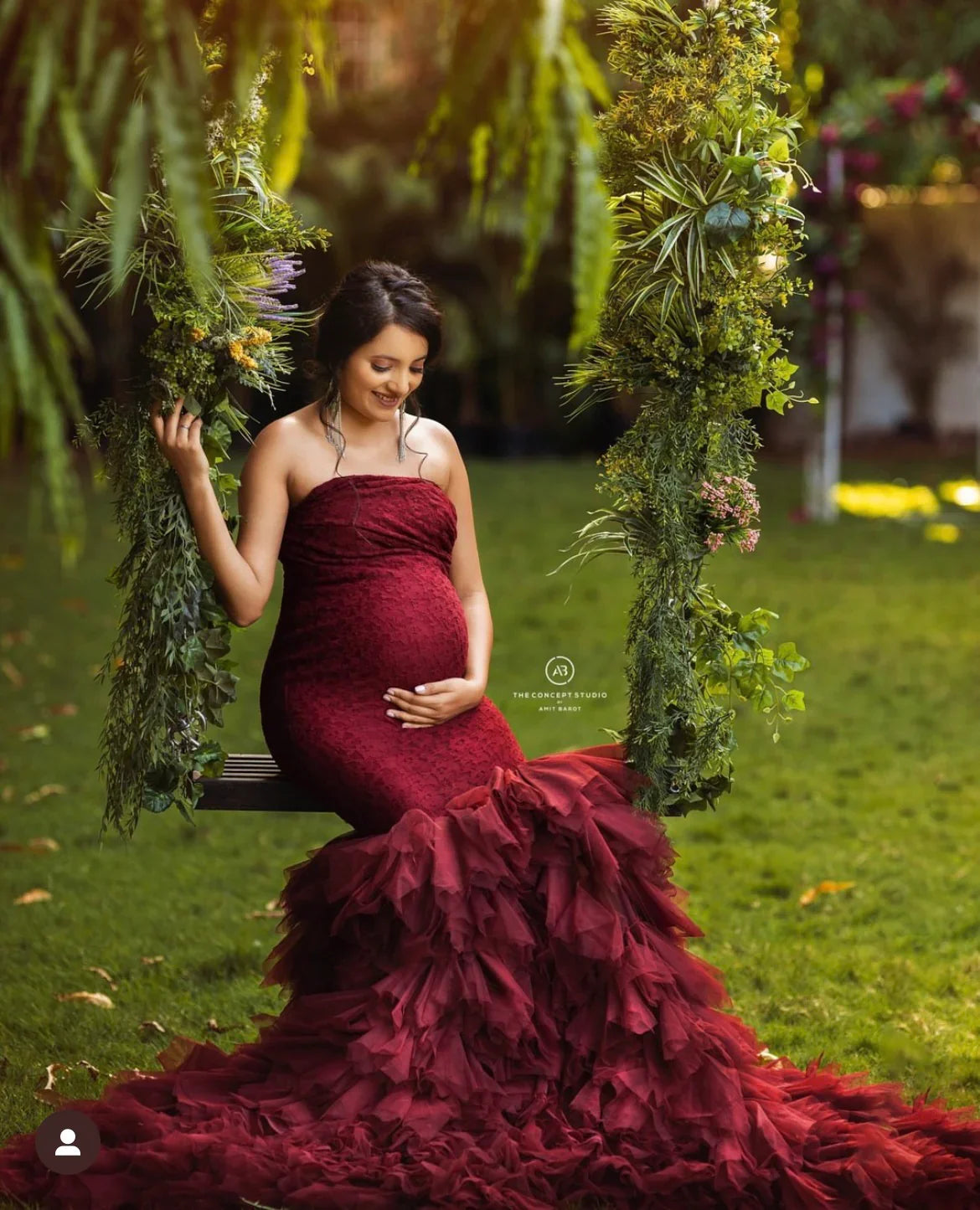 Pregnant woman in a red ruffled maternity gown sitting on a floral swing outdoors