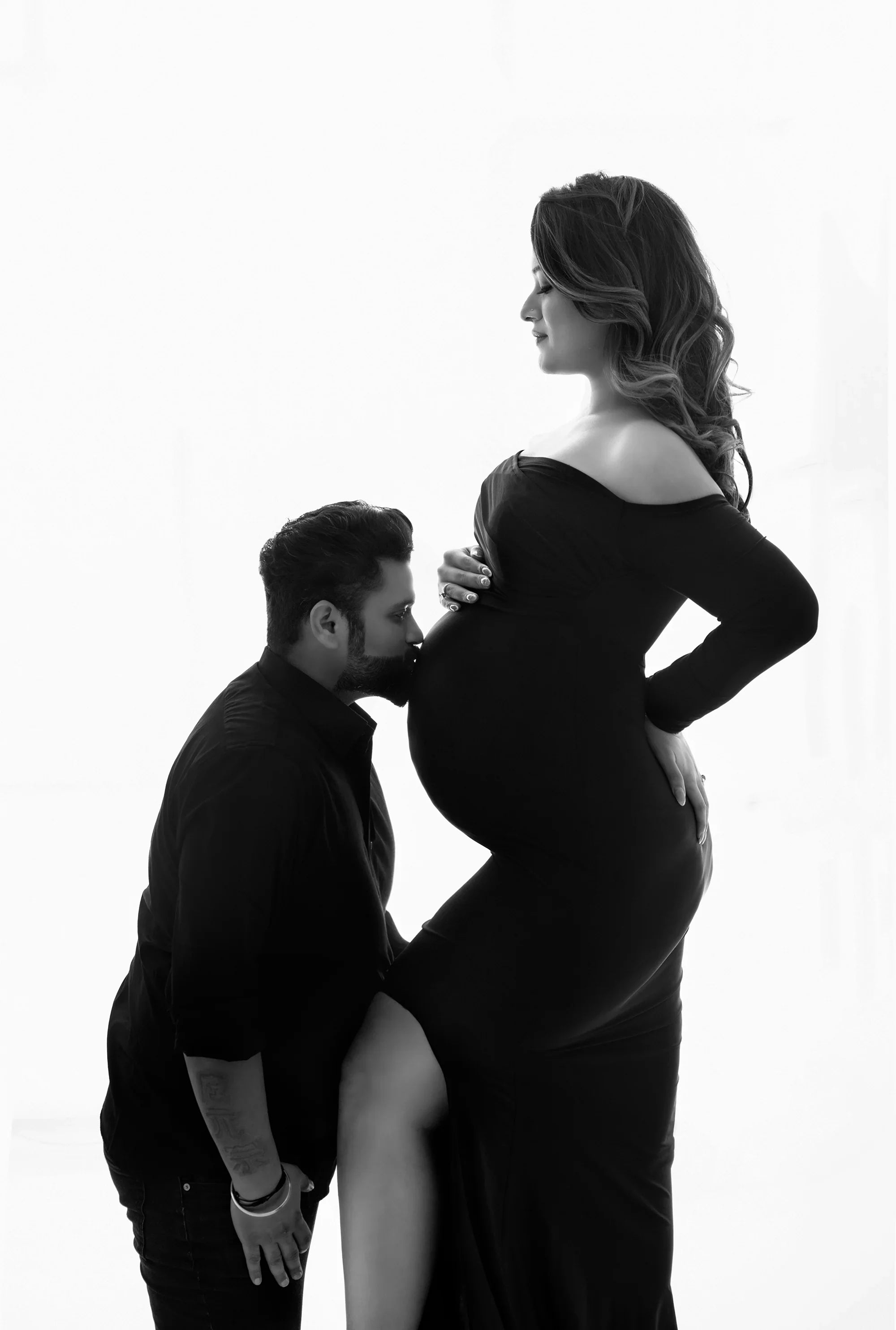 Maternity photoshoot with woman in elegant off-shoulder gown and partner kissing baby bump
