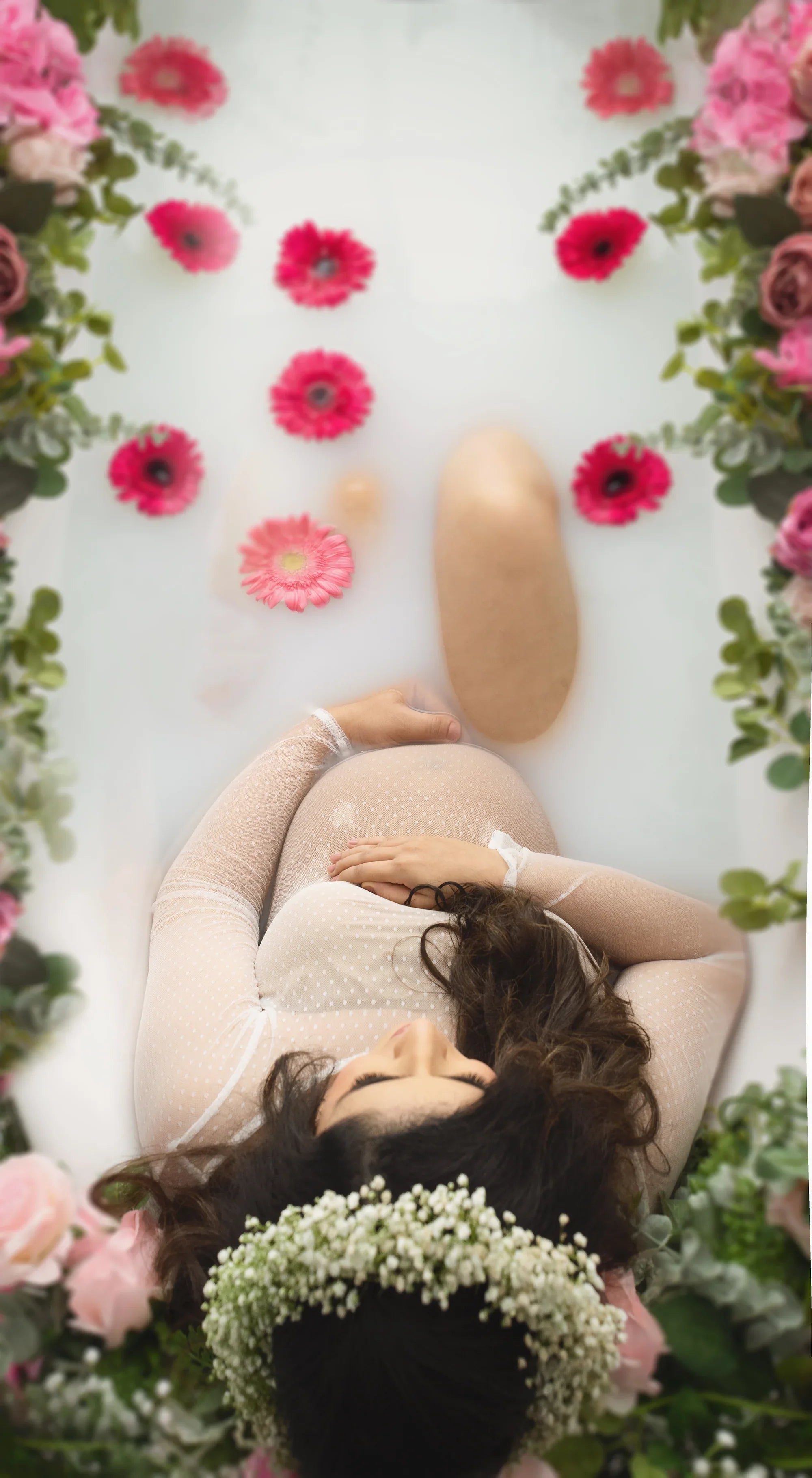 Pregnant woman in sheer dotted gown relaxing in a milk bath with pink flowers and greenery