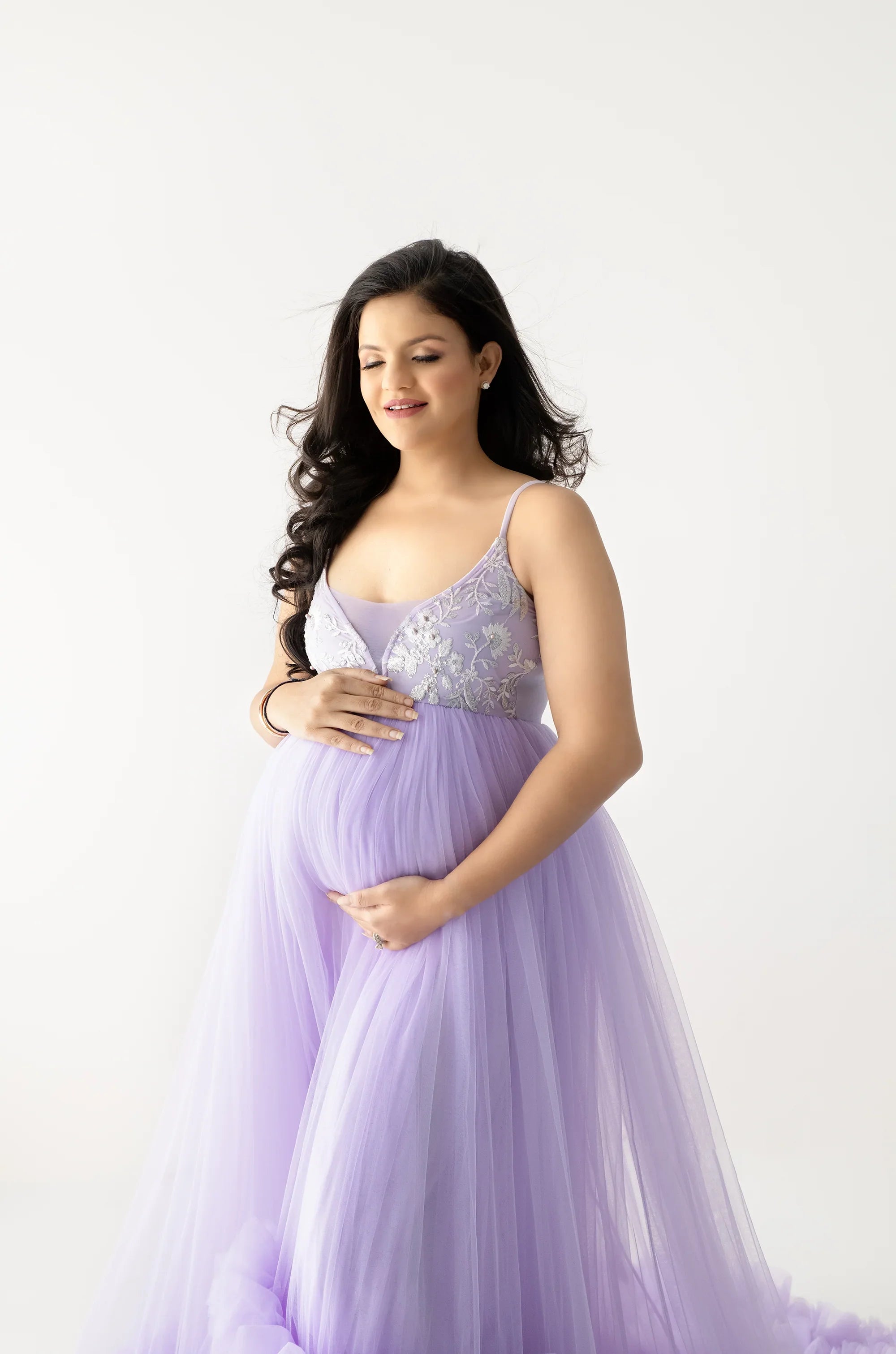 Pregnant woman in a lavender tulle maternity gown with floral embroidery, studio background.
