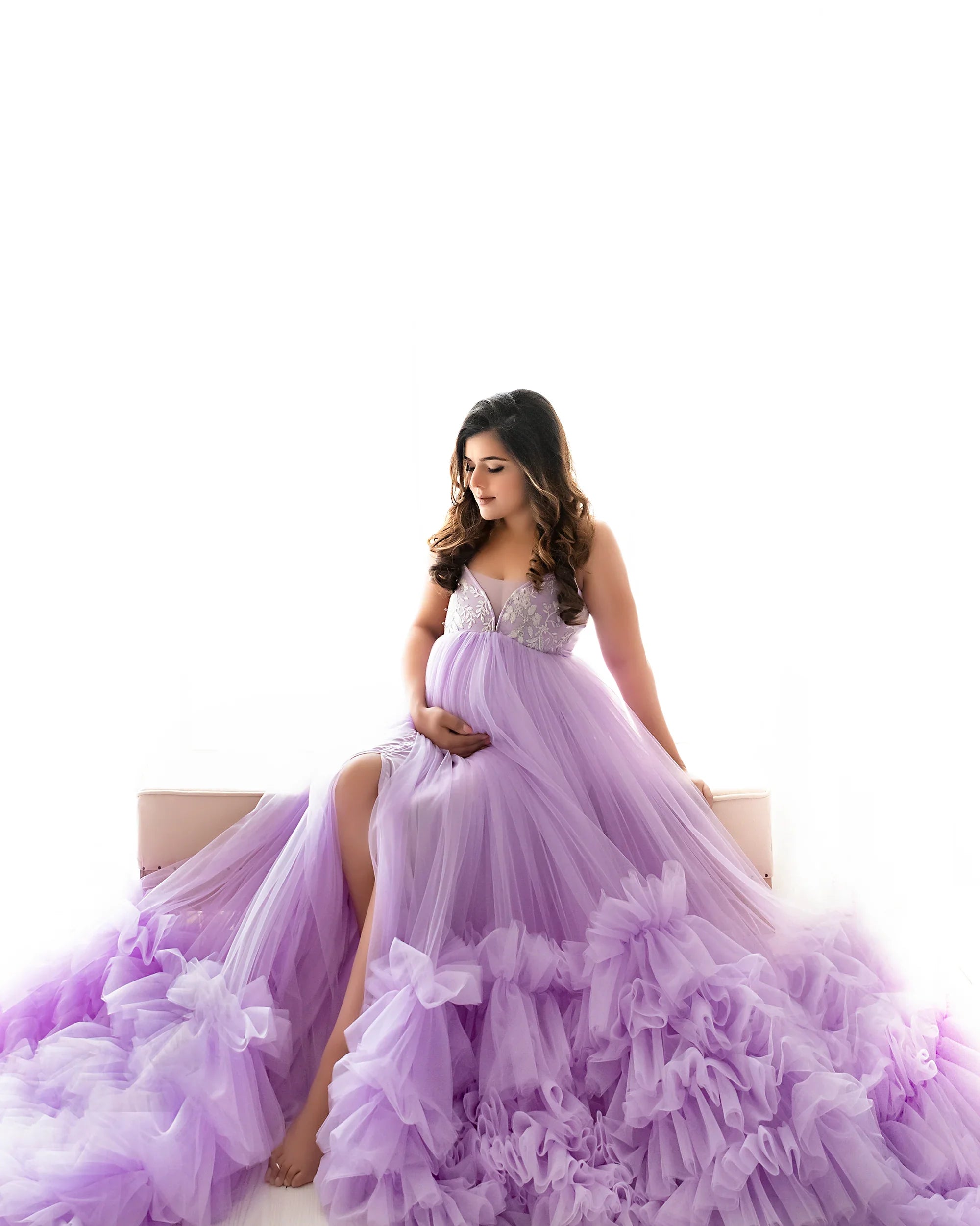 Pregnant woman in a flowing lavender maternity gown with ruffled tulle skirt, sitting indoors.