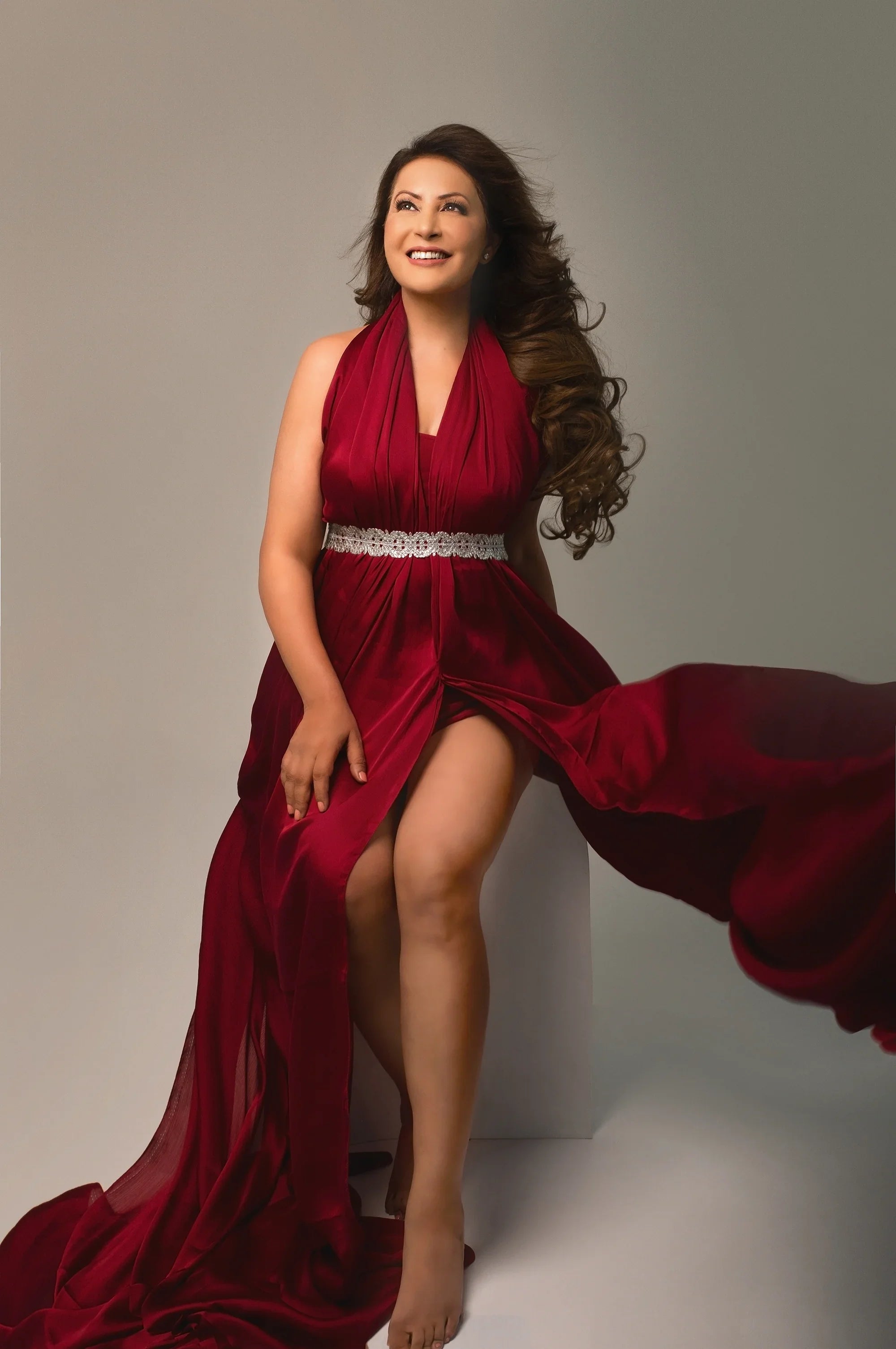 Smiling woman in a flowing burgundy maternity gown with lace belt, studio setting