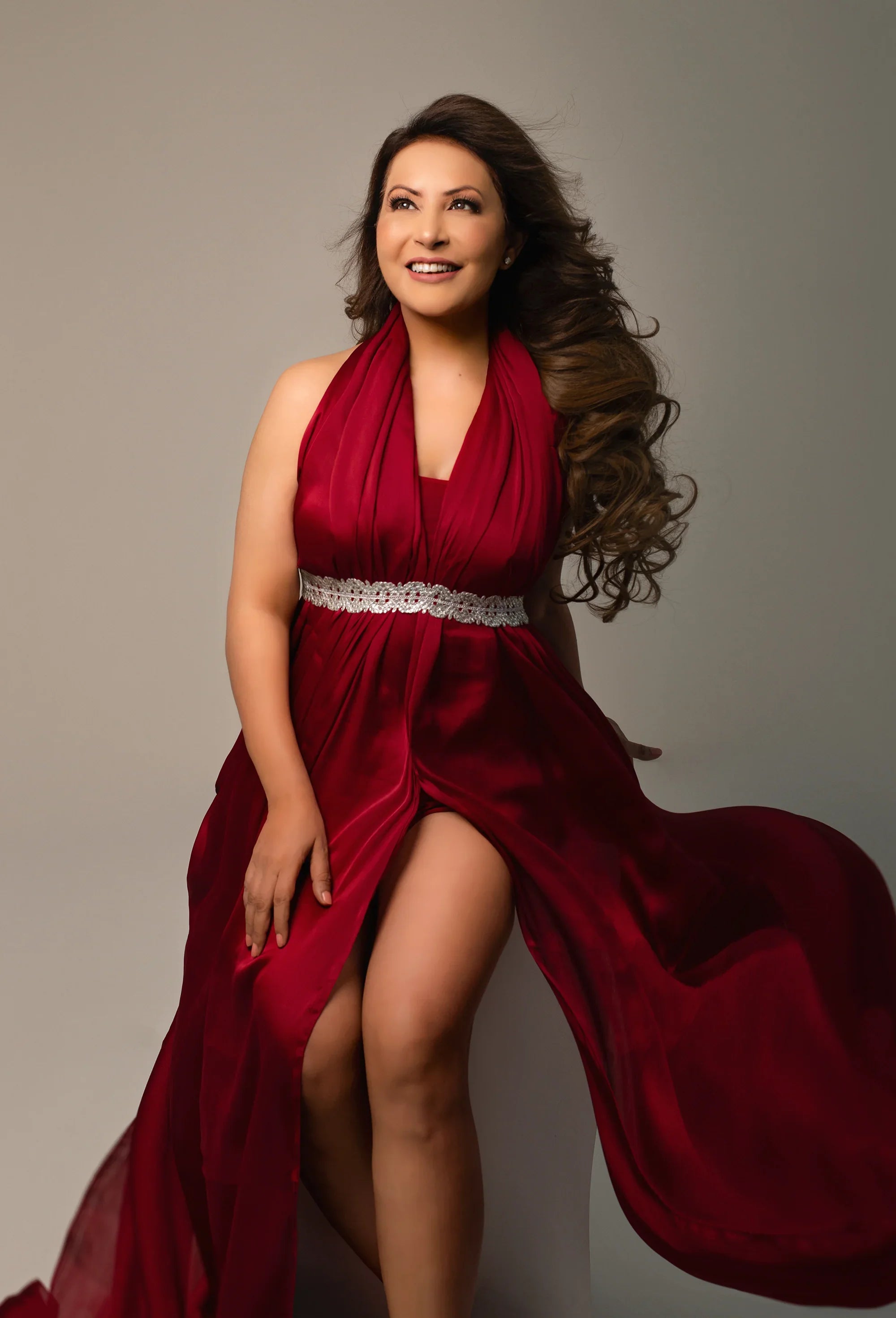 Woman in elegant red maternity gown with jeweled waist, smiling in studio setting