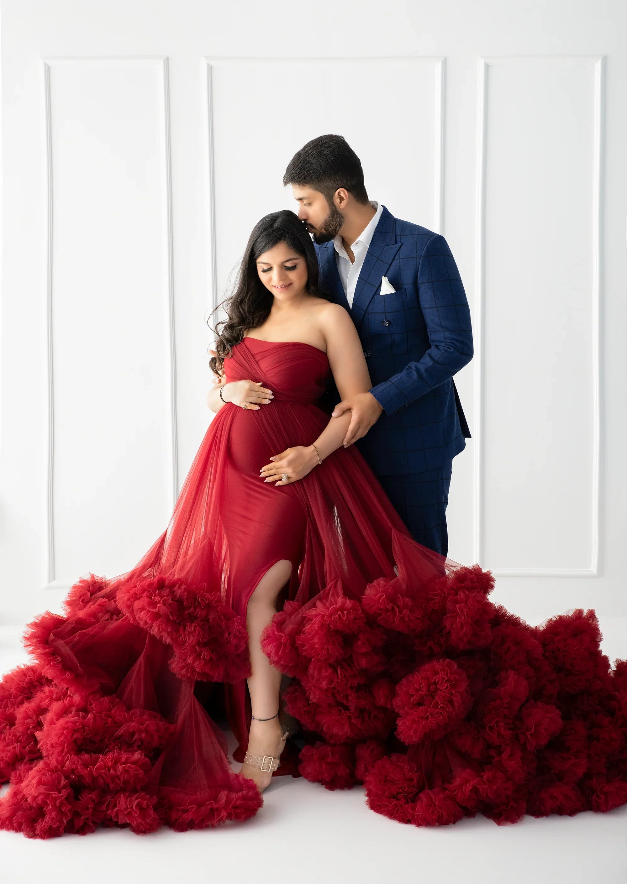 Pregnant woman in elegant red maternity gown posing with partner in blue suit, white background