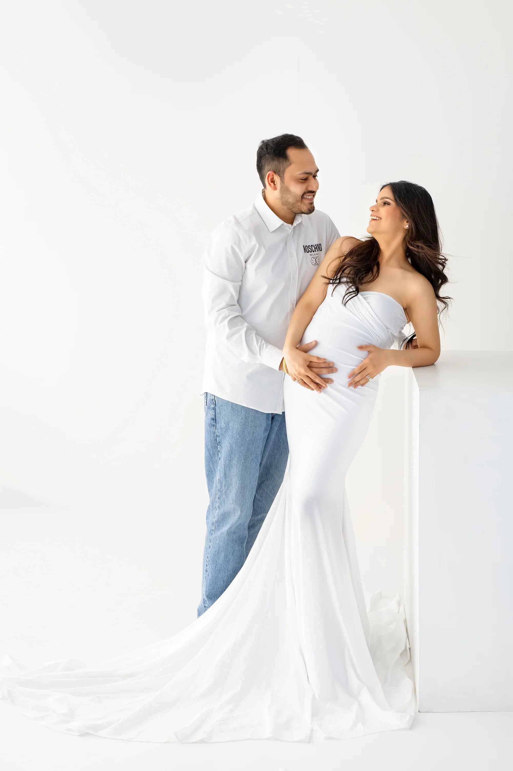 Smiling pregnant woman in an elegant white maternity gown with partner in a minimal studio
