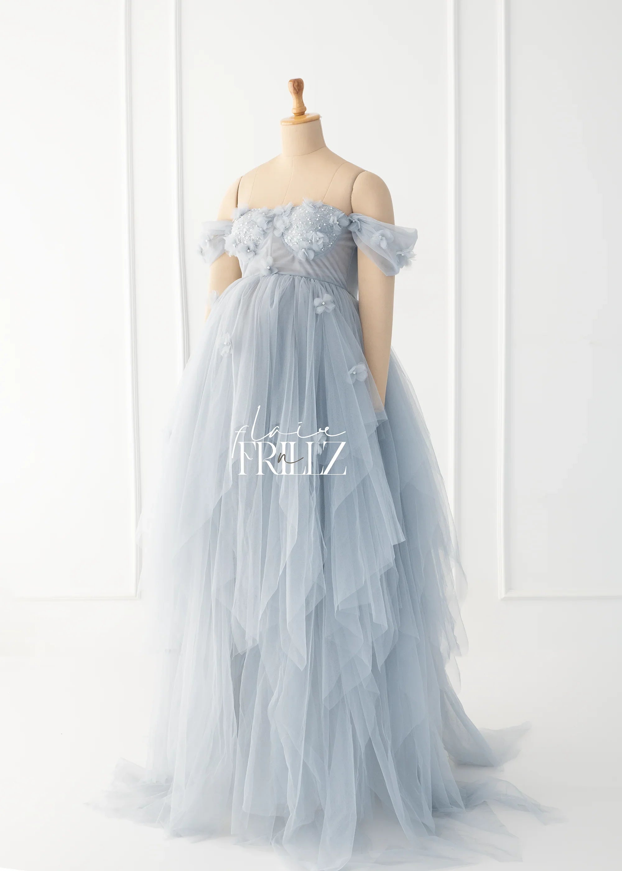 Light blue tulle maternity gown with floral appliqués on a mannequin in a bright studio.