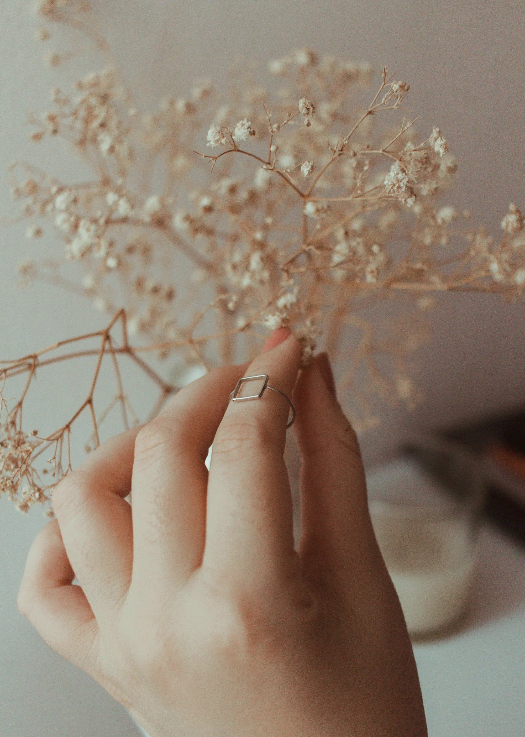Delicate hand with minimalist square ring gently touching dried baby's breath flowers, soft neutral tones.