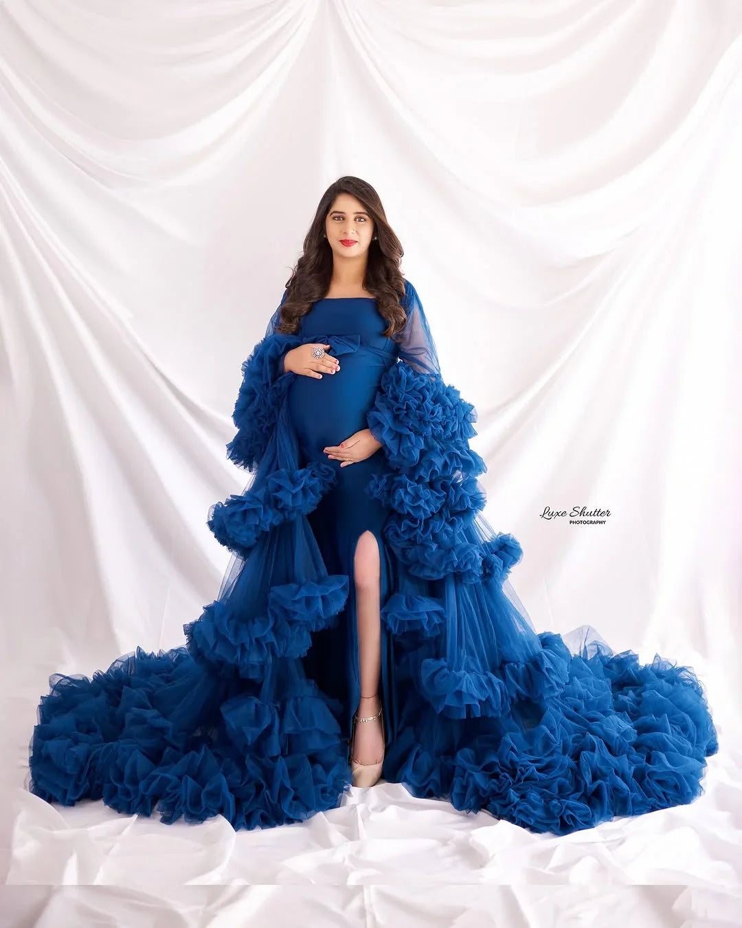 Pregnant woman in a royal blue ruffled maternity gown posing against a white draped backdrop