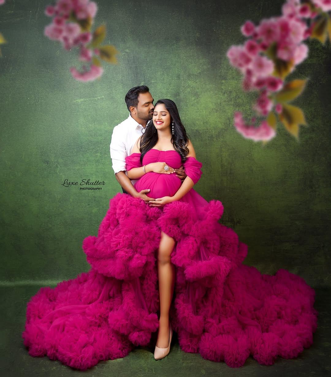 Pregnant woman in vibrant pink maternity gown embraced by partner, floral studio backdrop.