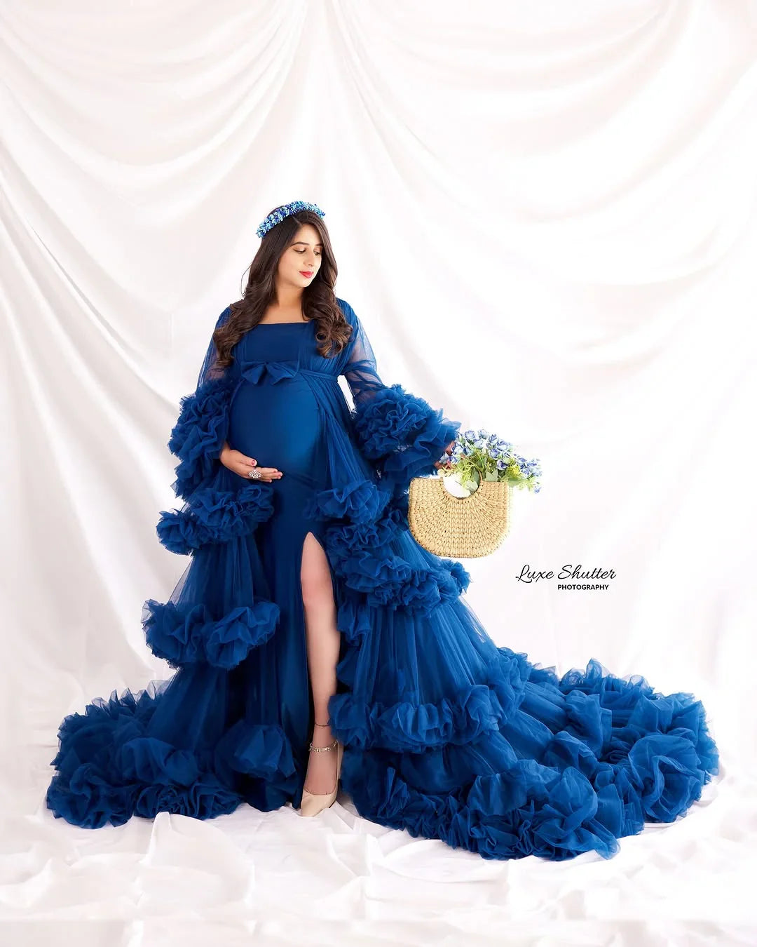 Pregnant woman in a flowing blue ruffle maternity gown with a floral headband, holding belly