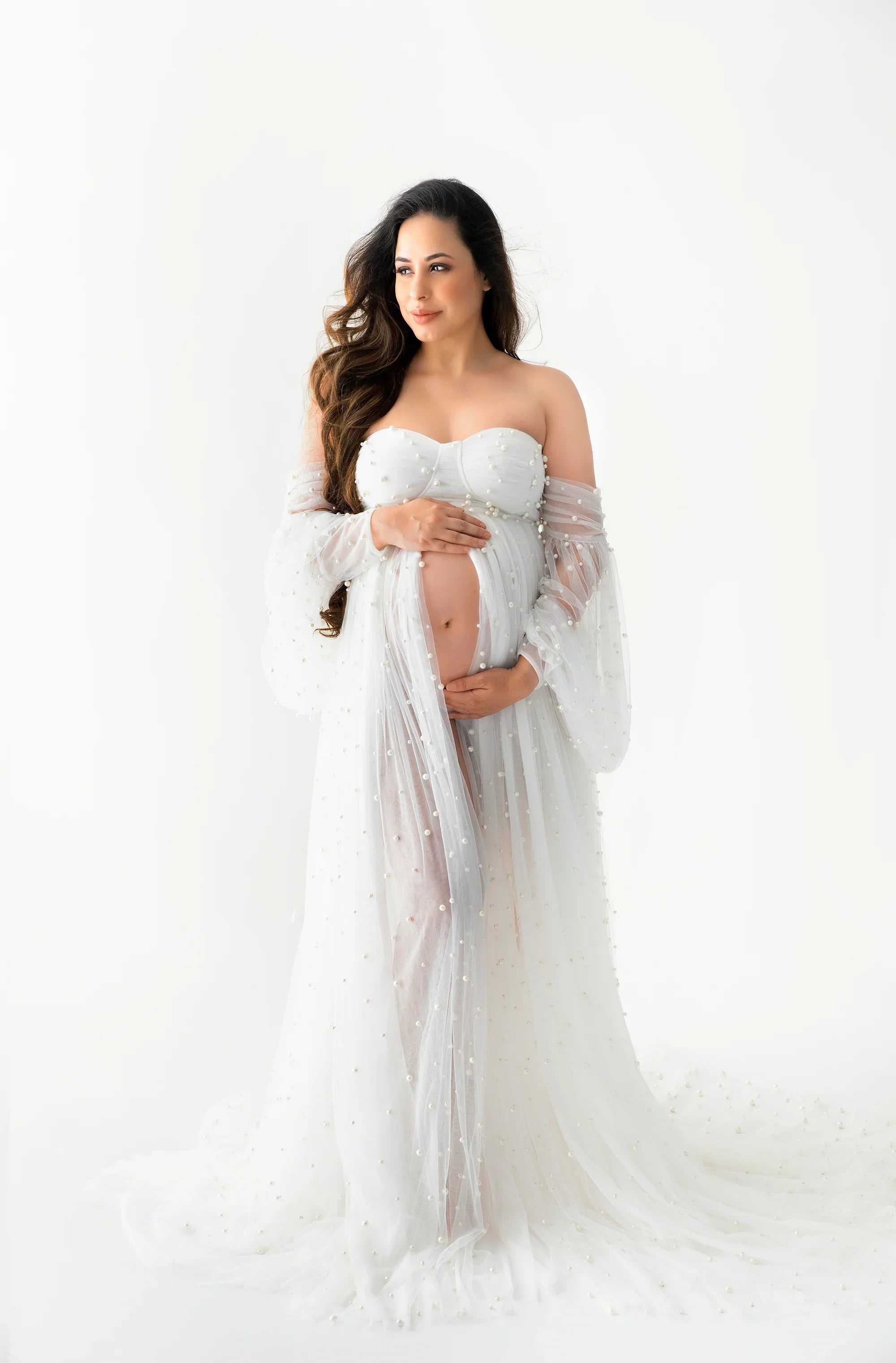 Pregnant woman in white sheer pearl embellished maternity gown posing against white background