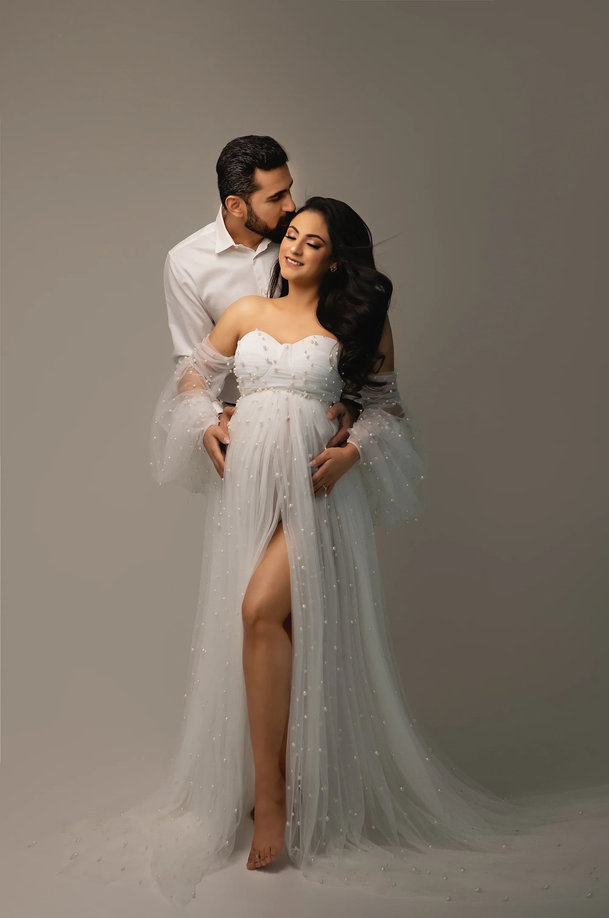 Couple posing in studio, pregnant woman in elegant white maternity gown with sheer pearl sleeves
