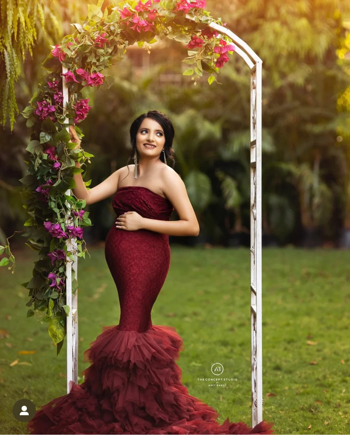 Pregnant woman in a burgundy maternity gown posing outdoors under a floral arch