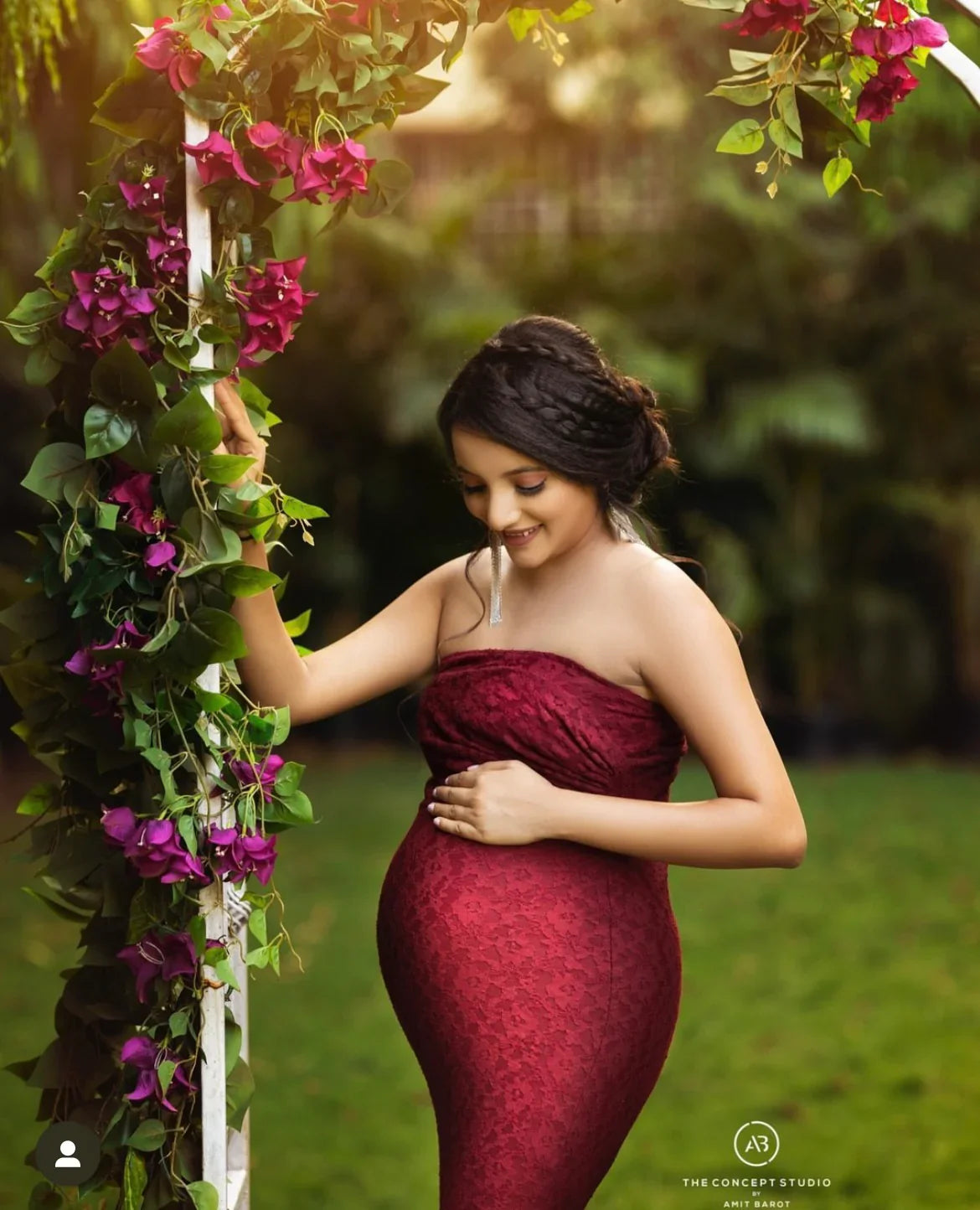 Pregnant woman in red lace maternity gown posing outdoors by floral arch