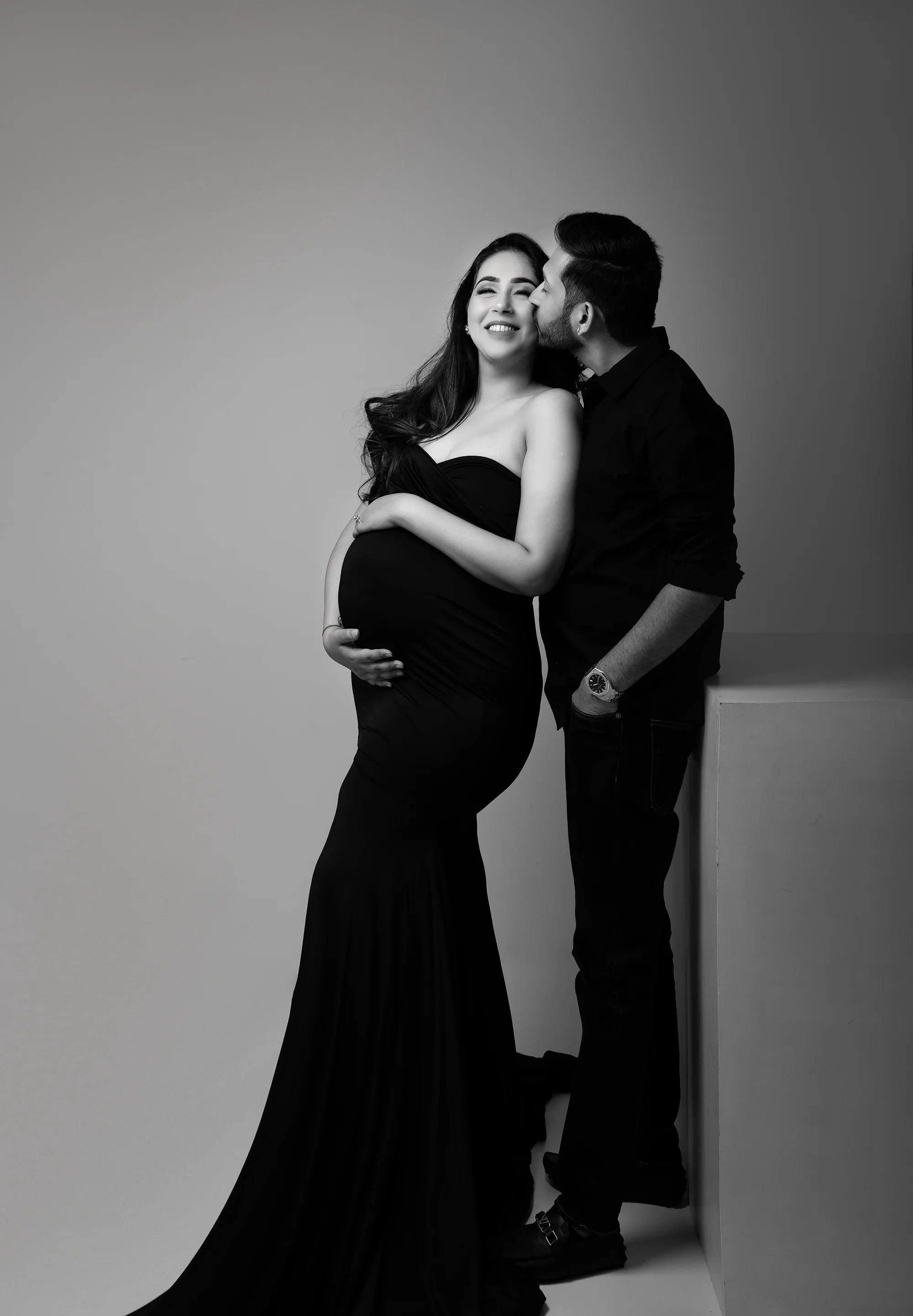 Smiling pregnant woman in elegant black maternity gown with partner kissing her forehead