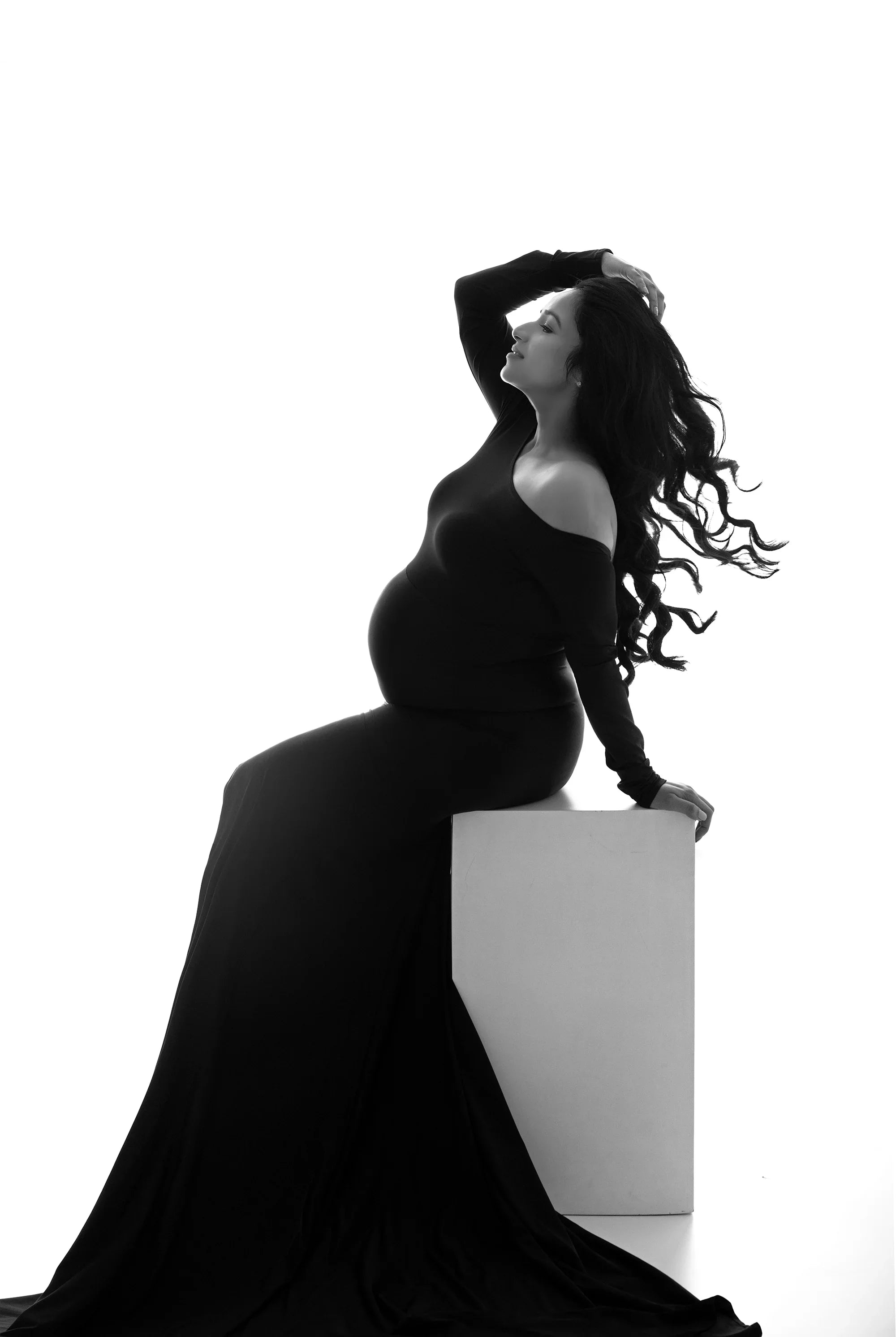 Pregnant woman in elegant black maternity gown with long train, sitting on a white cube