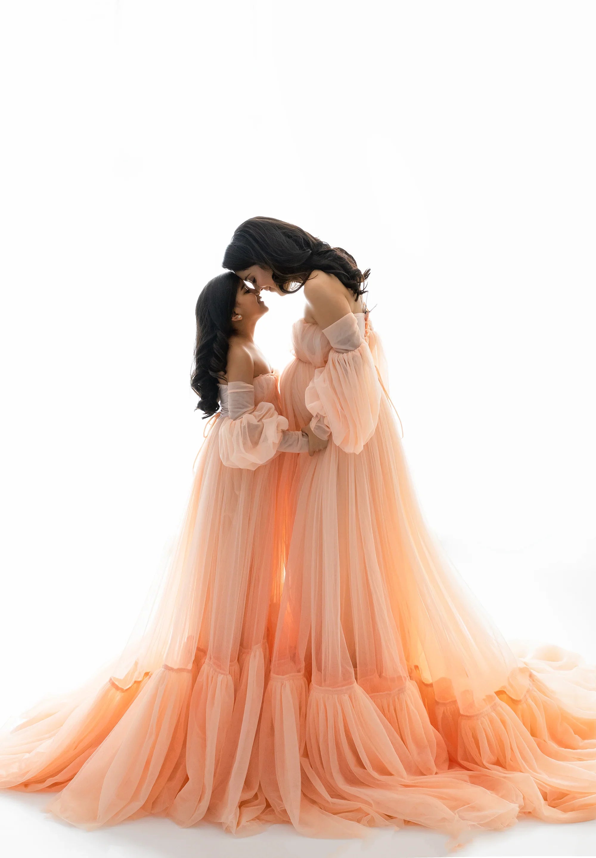 Pregnant woman and young girl in matching peach tulle gowns, embracing in a bright studio