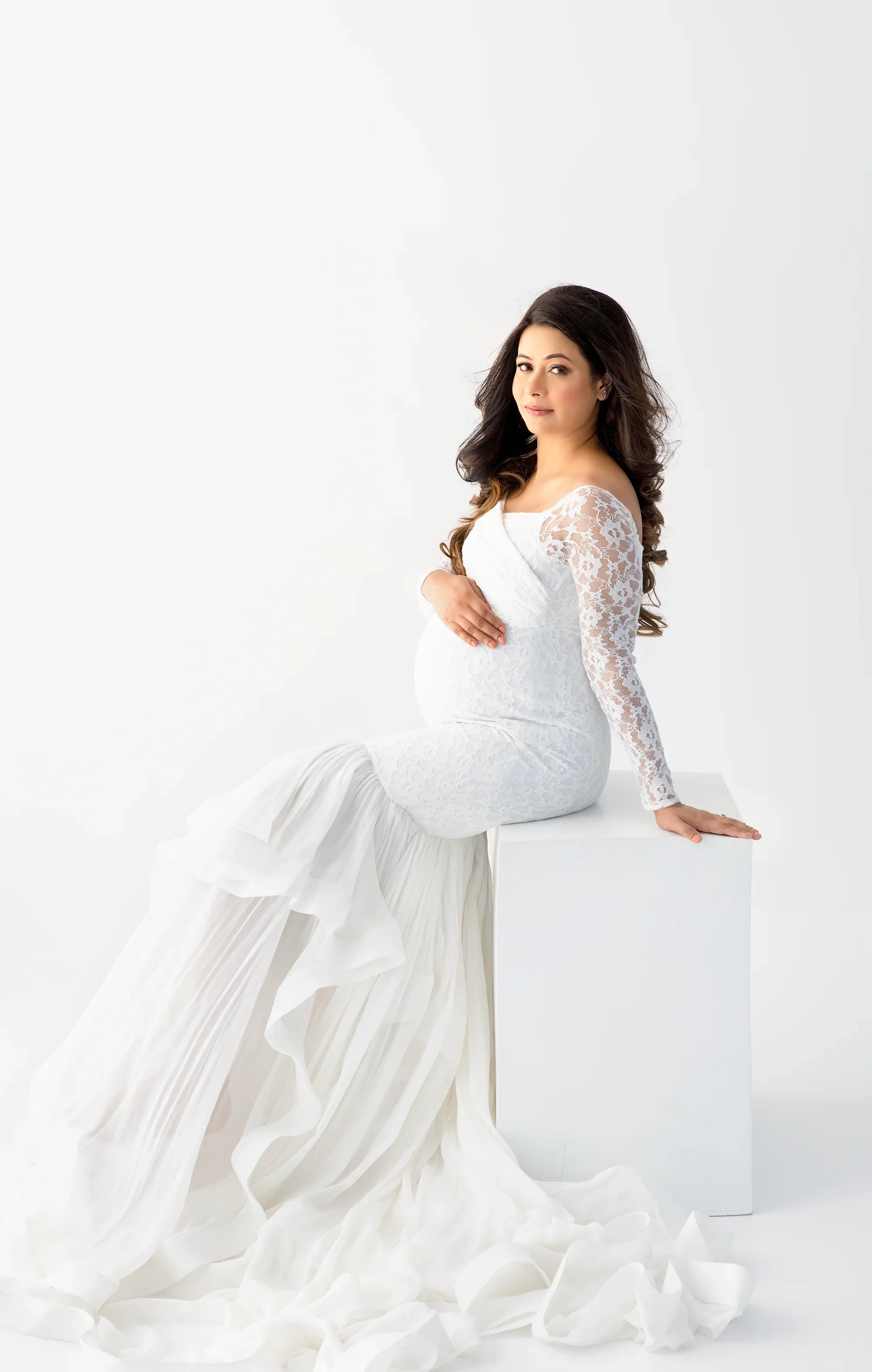 Pregnant woman in elegant white lace maternity gown with flowing skirt, posing on white cube