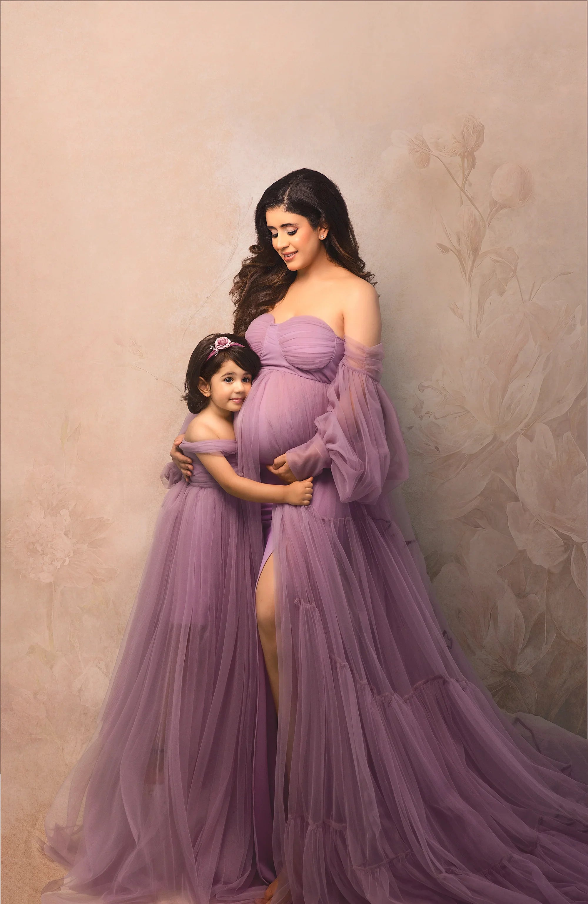 Pregnant woman and young girl in matching lavender maternity gowns against a floral backdrop