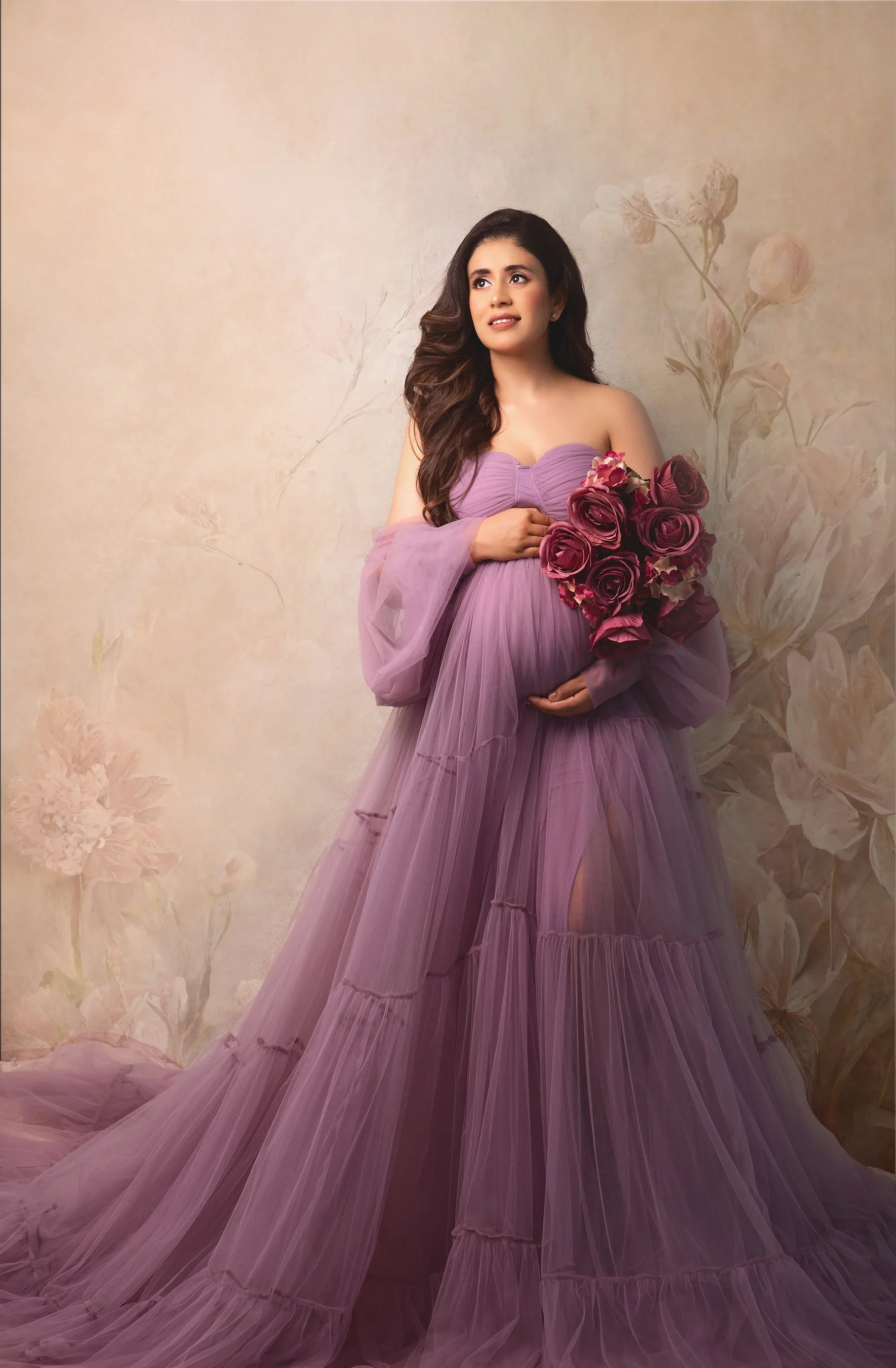 Pregnant woman in flowing lavender maternity gown holding bouquet of roses, elegant floral backdrop