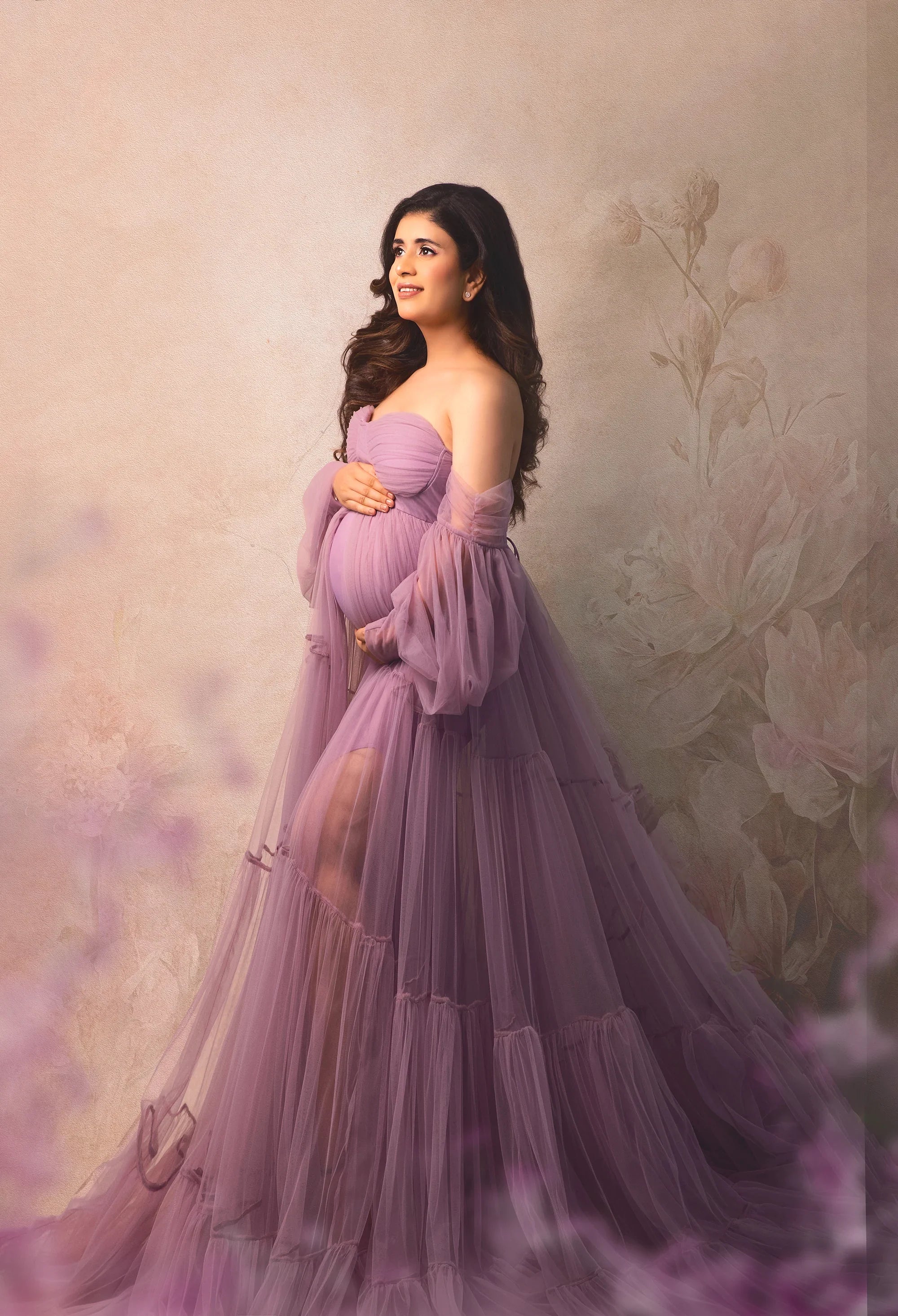 Pregnant woman in flowing lavender maternity gown posing against a floral backdrop