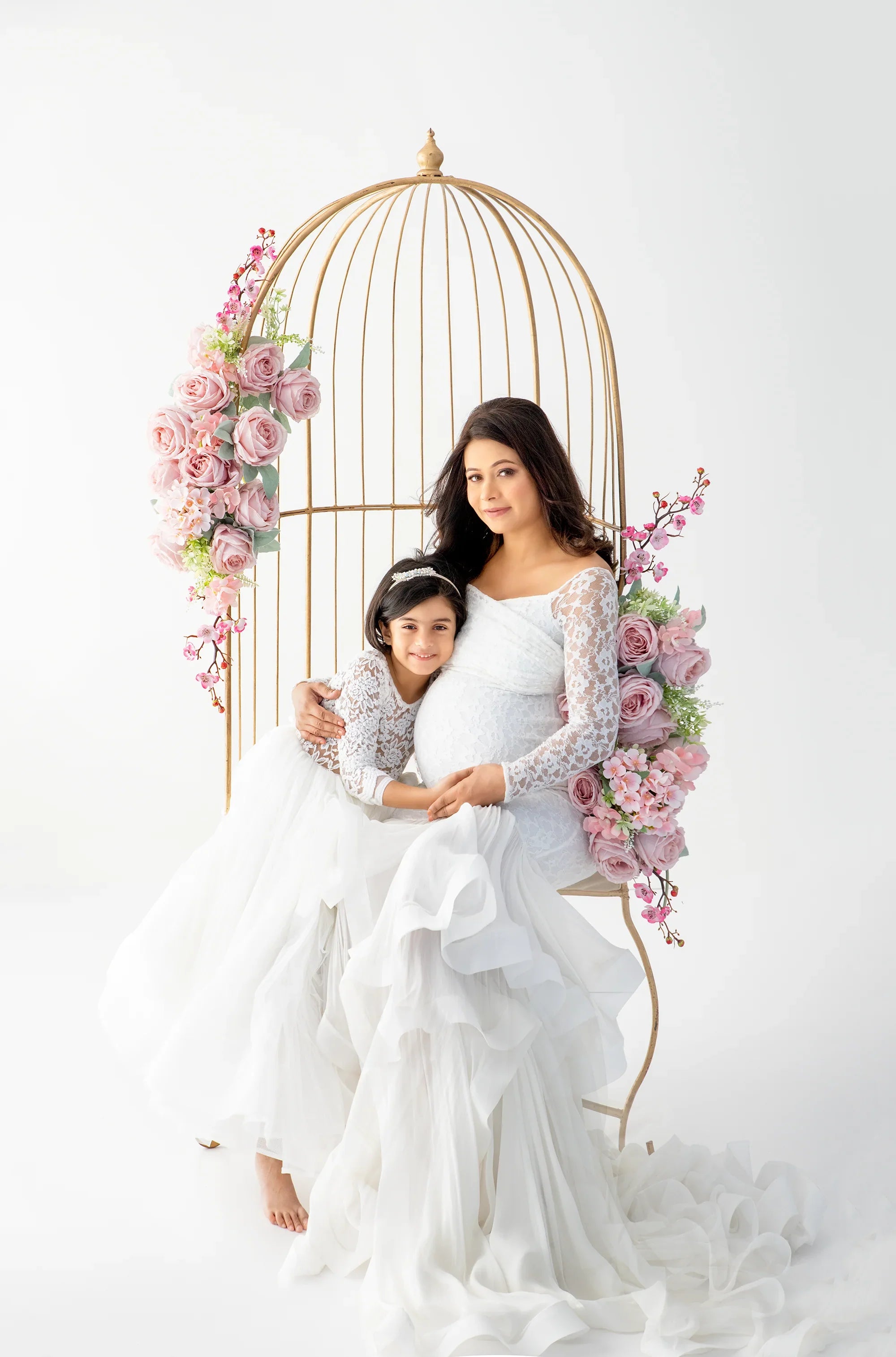 Pregnant woman and girl in white lace maternity gowns sitting on floral birdcage chair