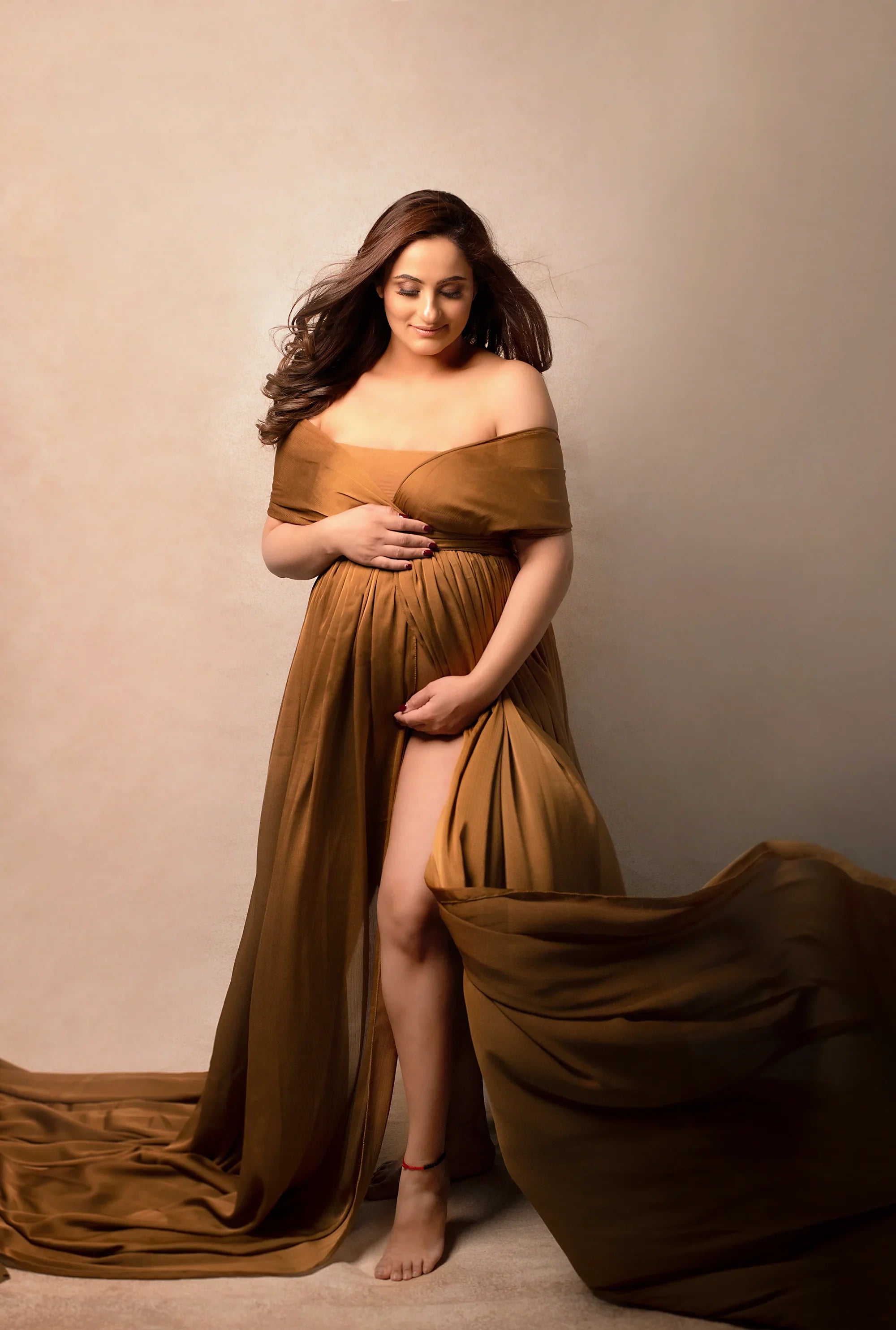 Pregnant woman in flowing brown off-shoulder maternity gown, posing in studio.