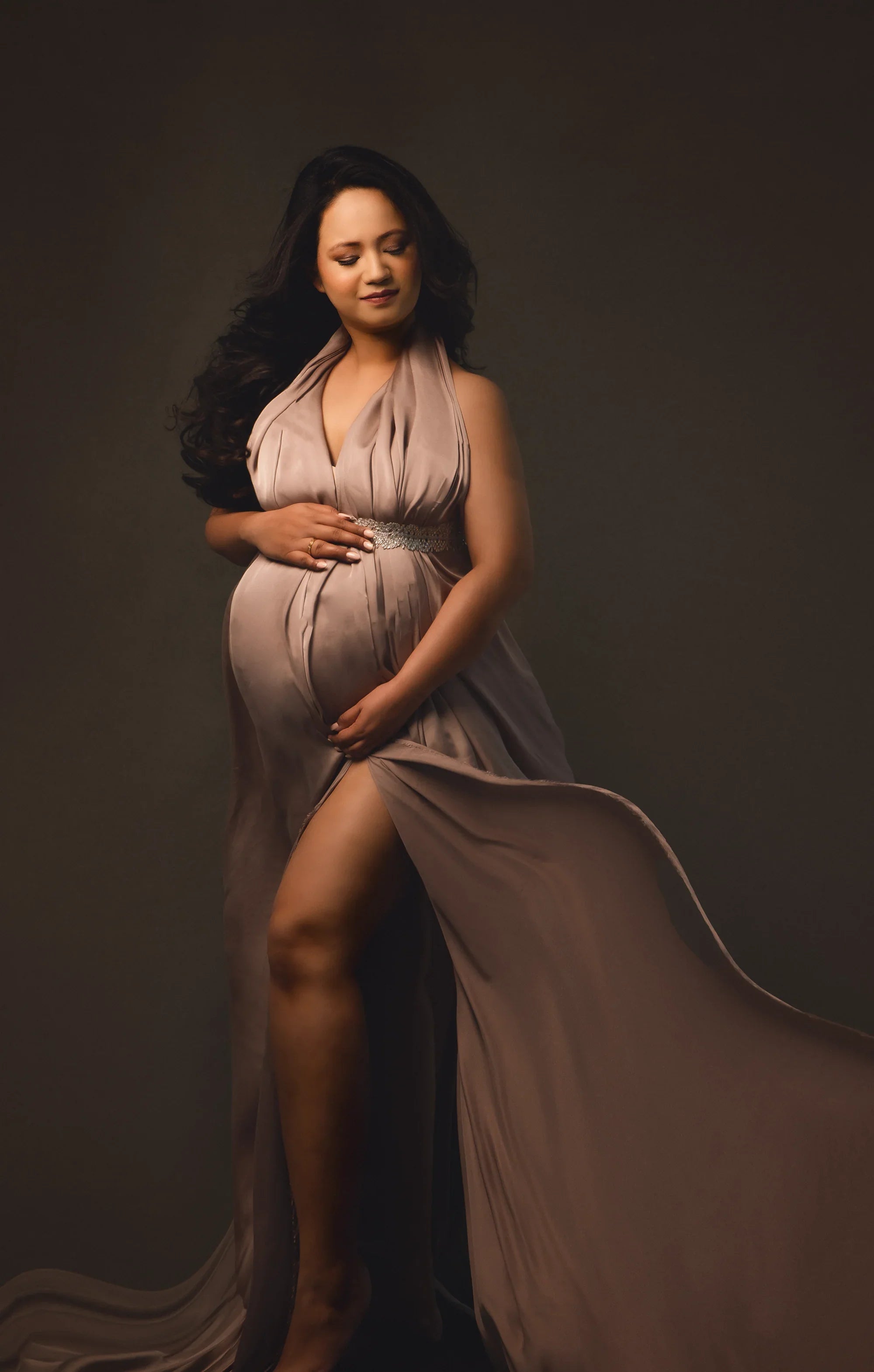 Pregnant woman in elegant taupe maternity gown posing against dark background