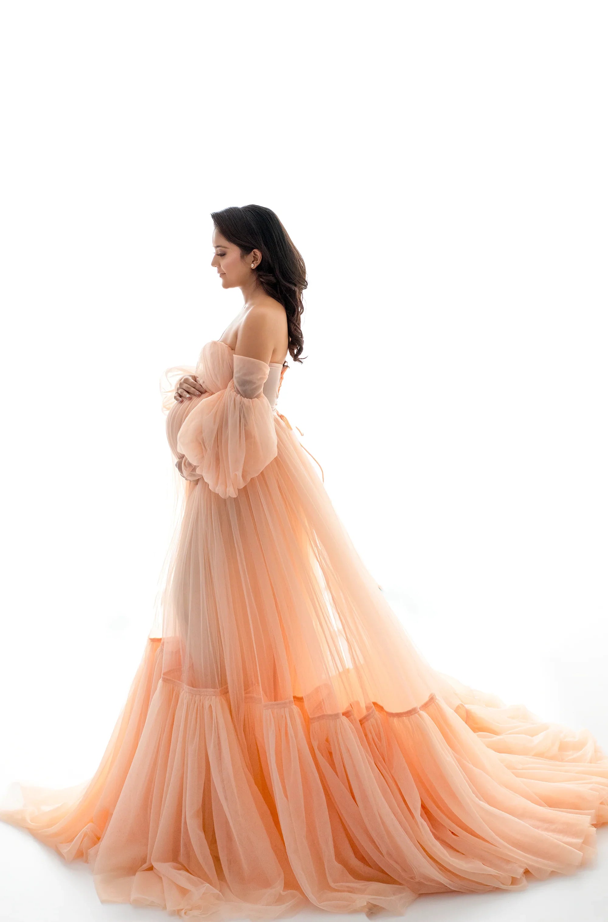 Pregnant woman in peach off-shoulder maternity gown with tulle skirt on white background