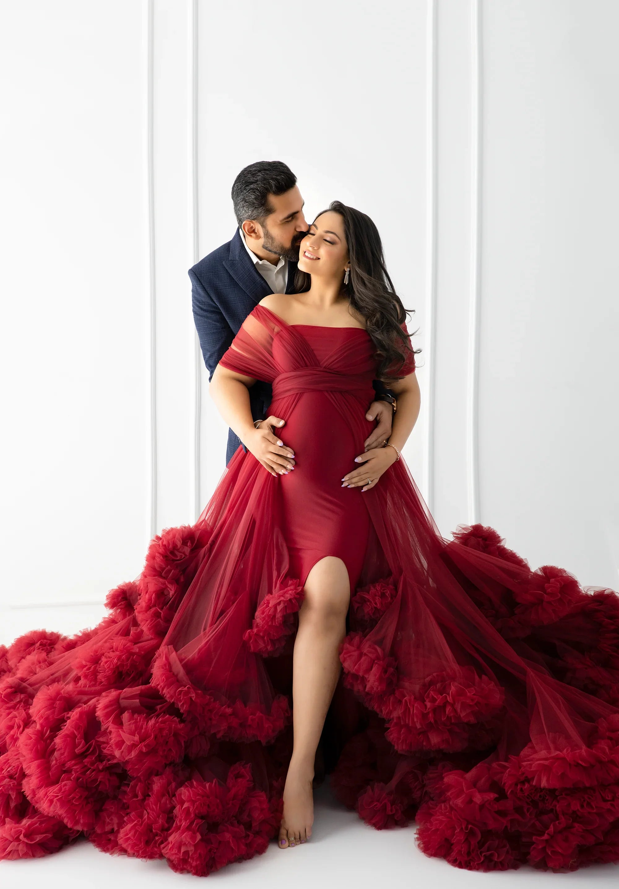 Pregnant woman in elegant red maternity gown with tulle, embraced by partner in suit, white studio