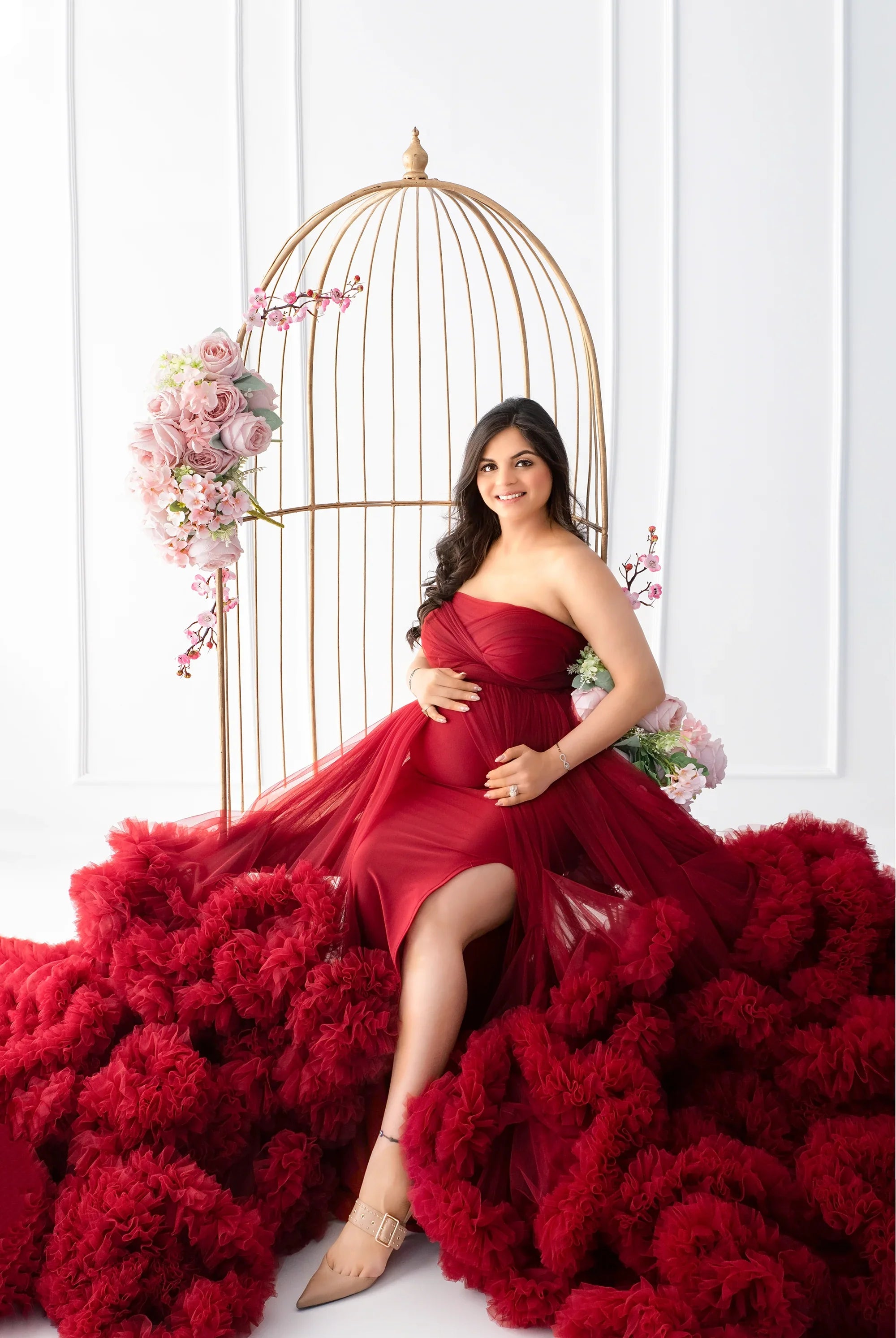 Pregnant woman in elegant red maternity gown with tulle train, sitting by floral birdcage