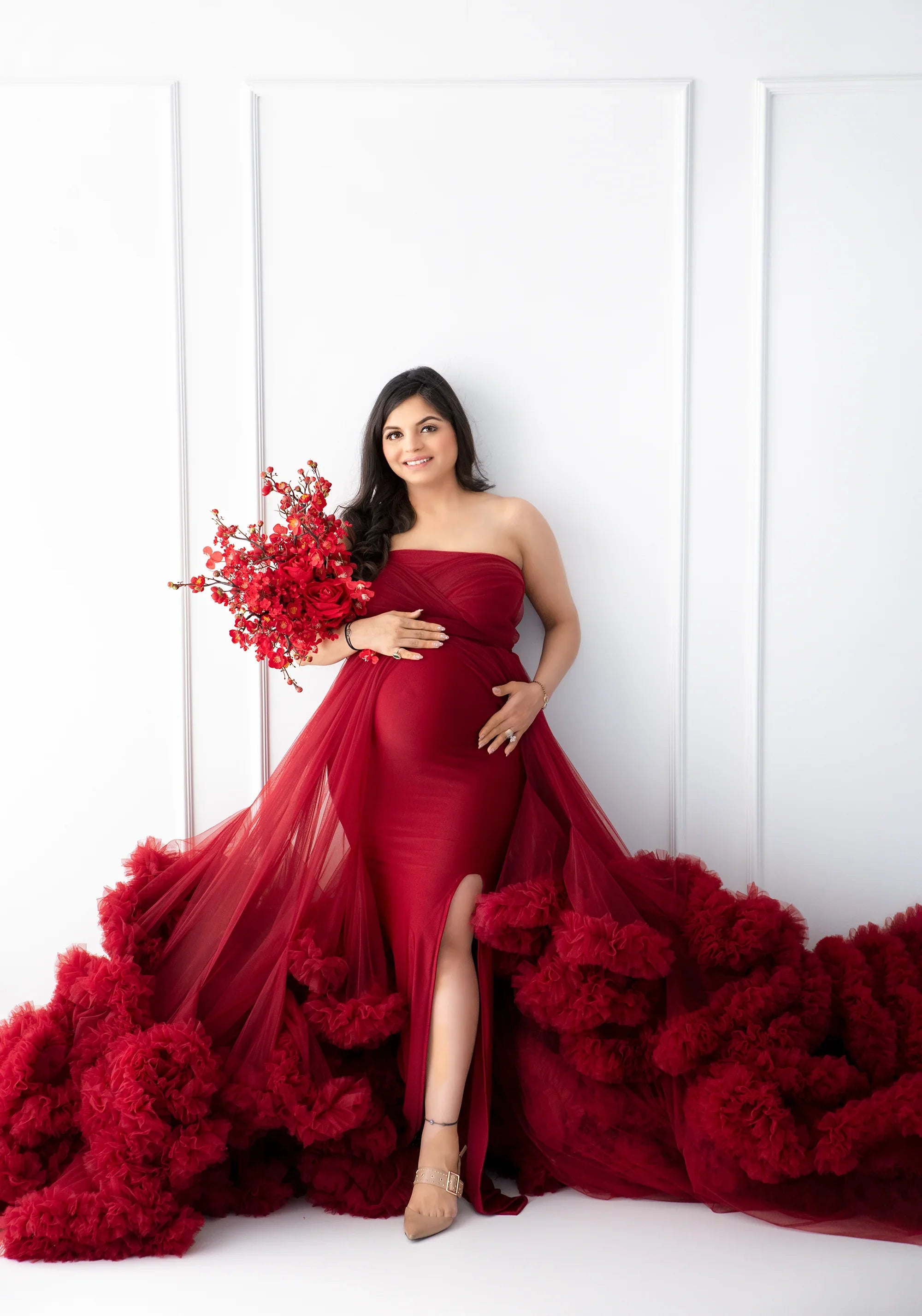 Pregnant woman in elegant red maternity gown holding red floral bouquet, white background