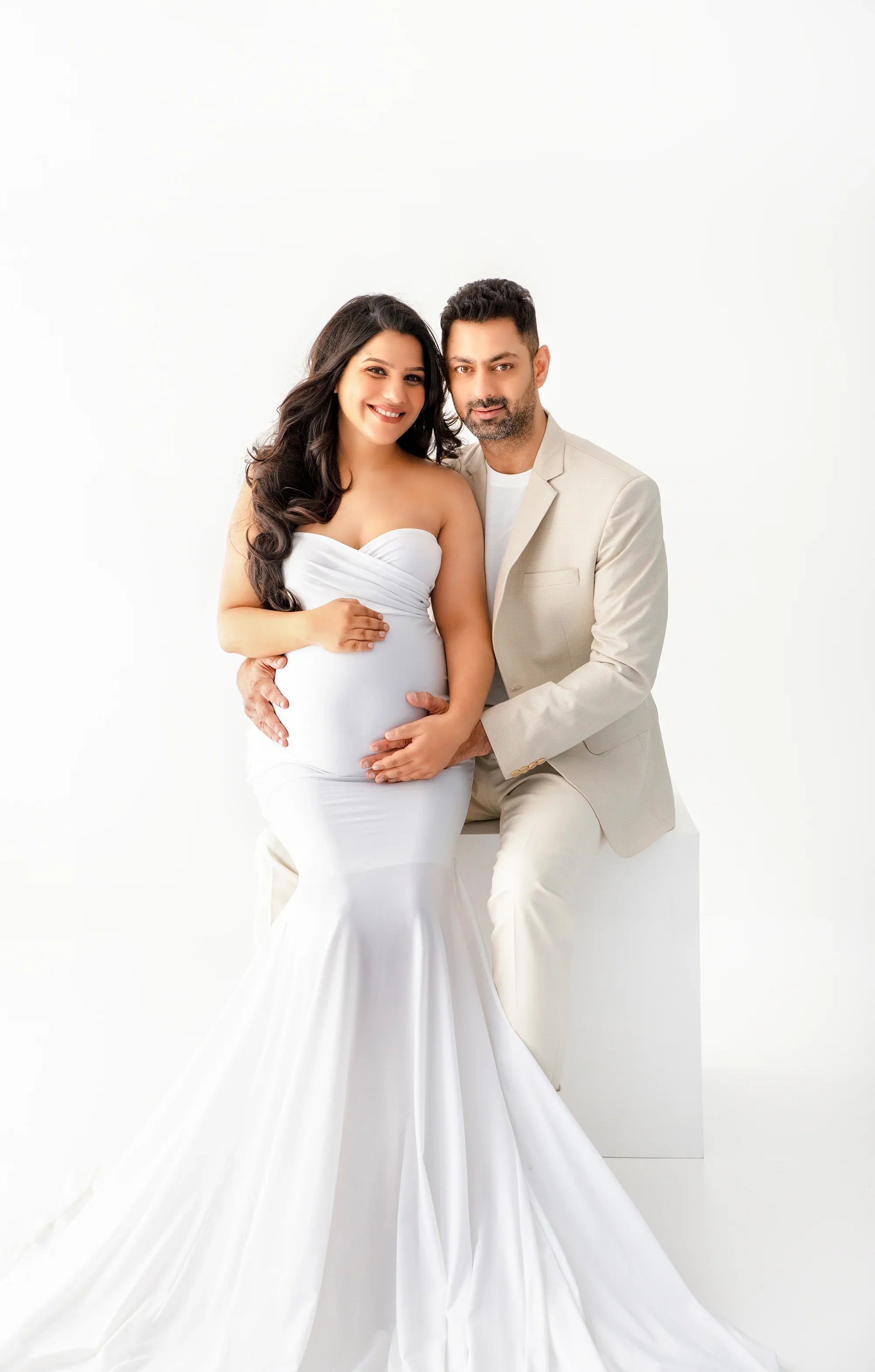 Smiling pregnant woman in elegant white maternity gown with partner in neutral suit, studio setting