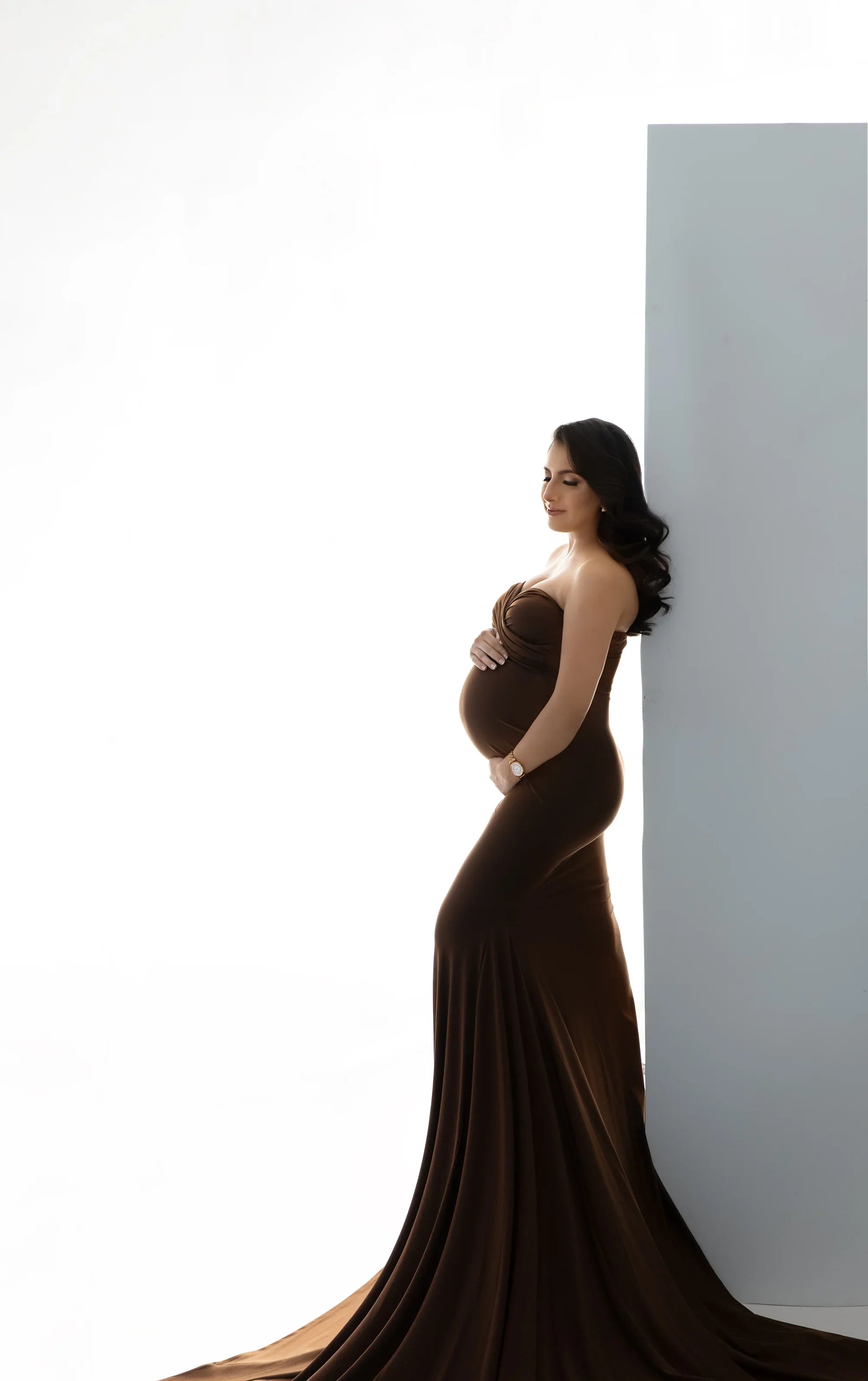 Pregnant woman in elegant brown strapless maternity gown posing against a light background.
