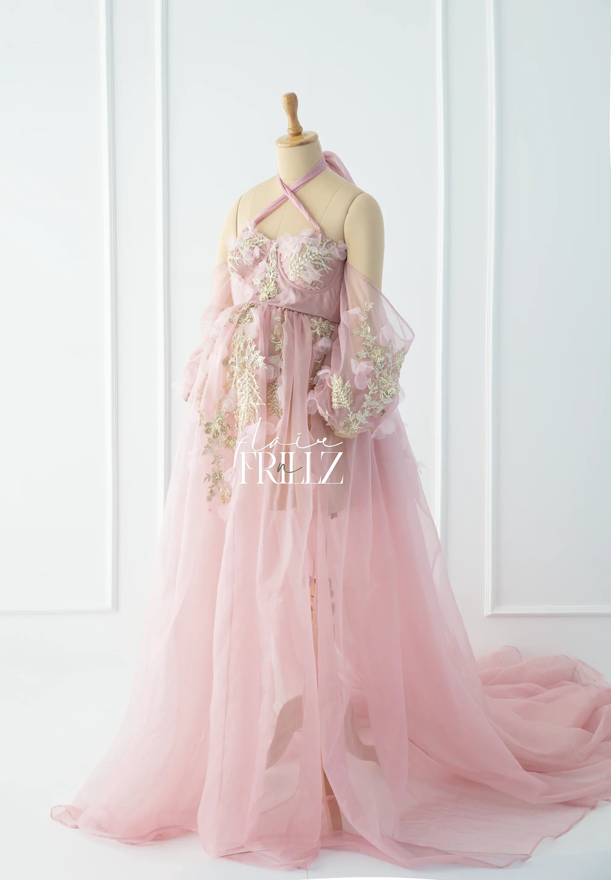 Pink maternity gown with sheer fabric, floral applique details, and off-shoulder sleeves on a mannequin