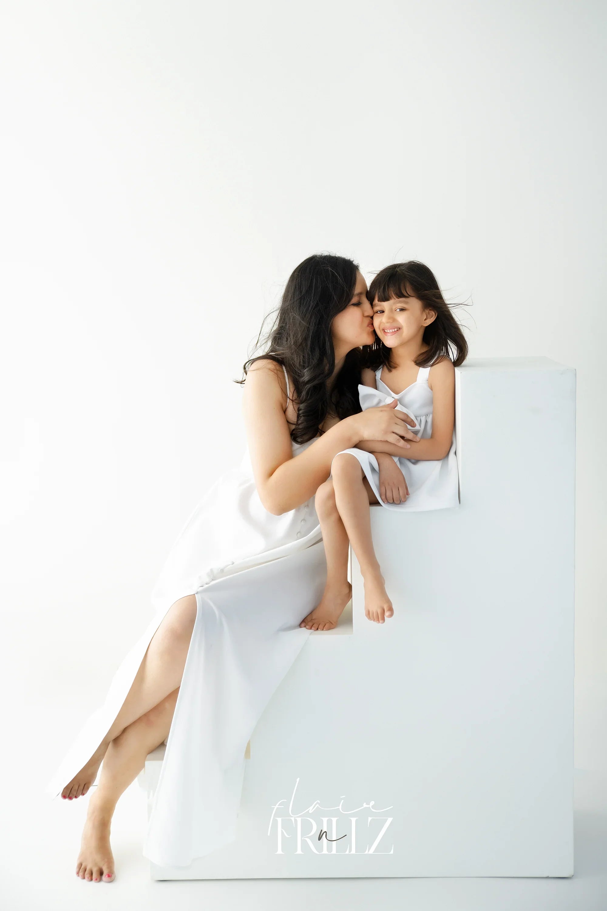 Mother in elegant white maternity gown kissing smiling daughter in matching dress, studio setting