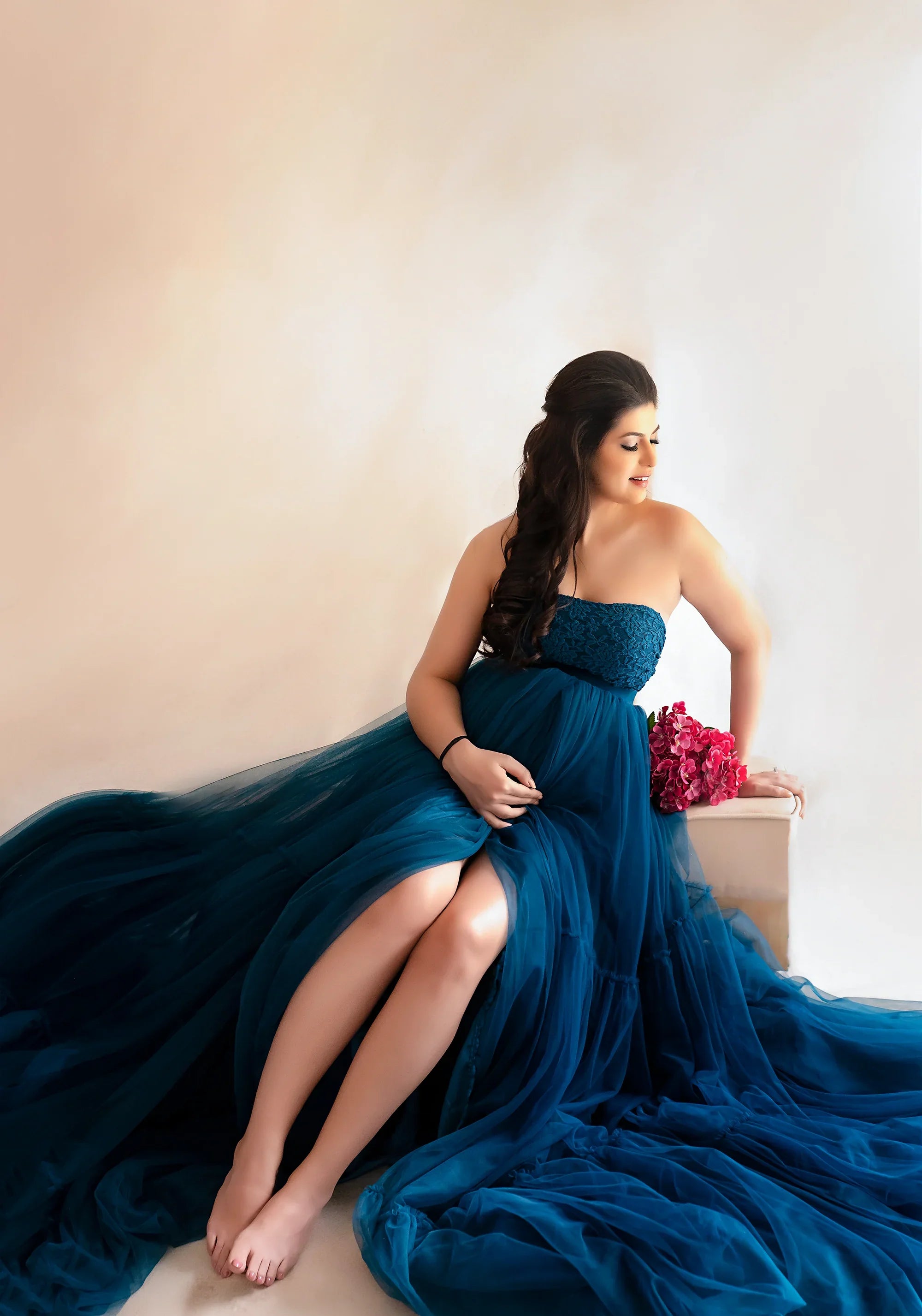 Pregnant woman in elegant blue maternity gown sitting indoors with pink flowers