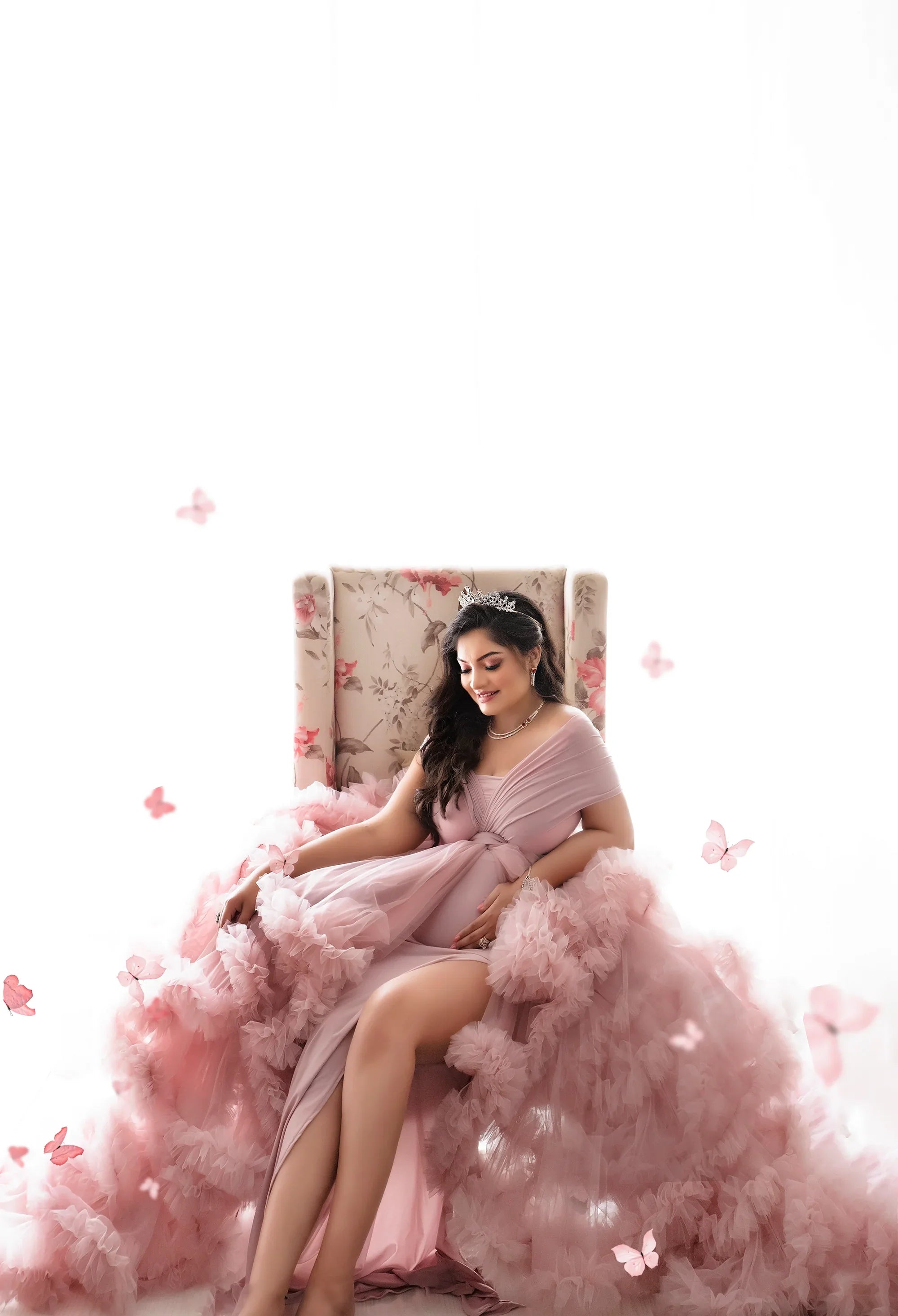 Pregnant woman in a pink ruffle maternity gown sitting on a floral chair with butterfly accents.
