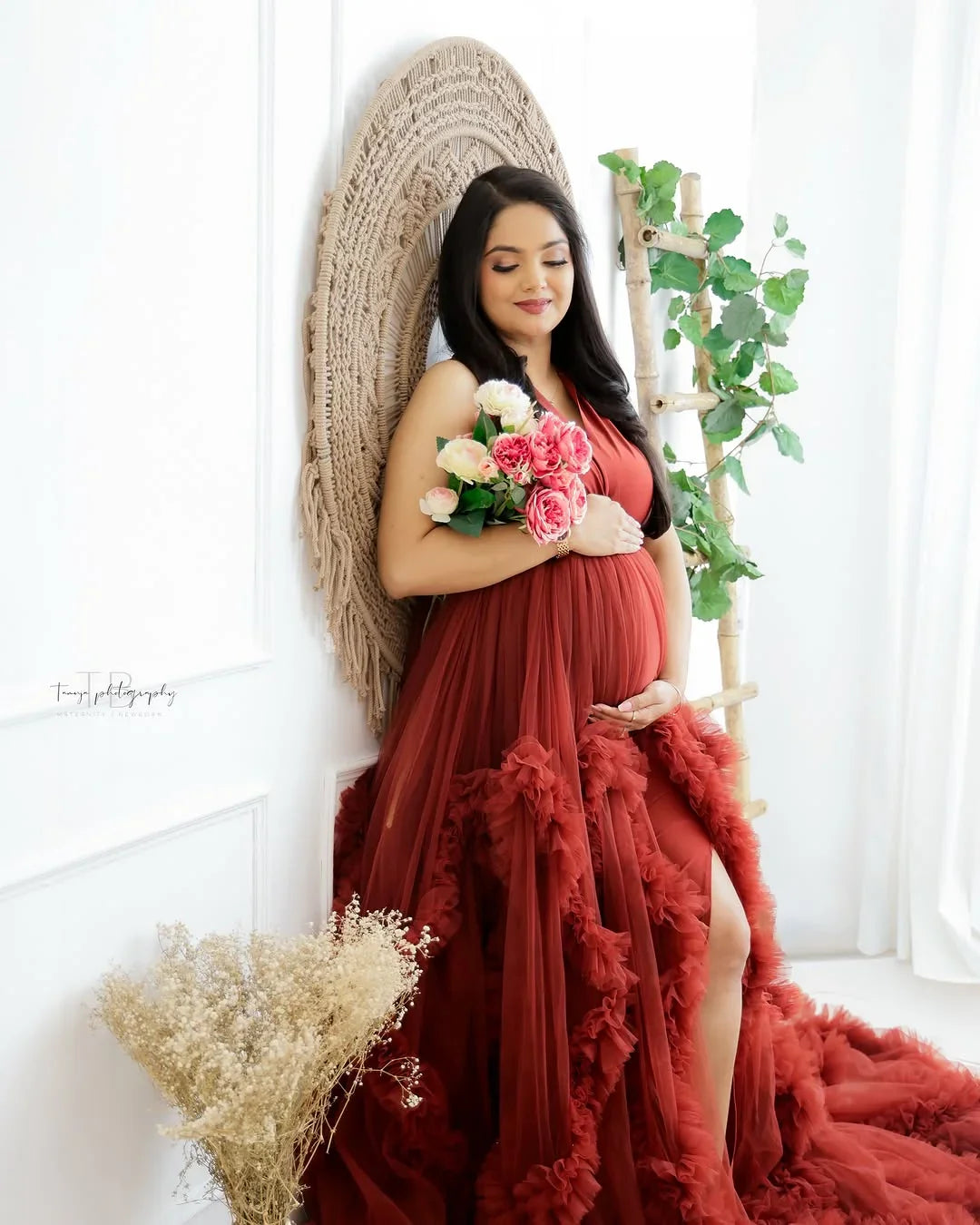 Pregnant woman in a flowing red maternity gown holding a bouquet in a bright, elegant room