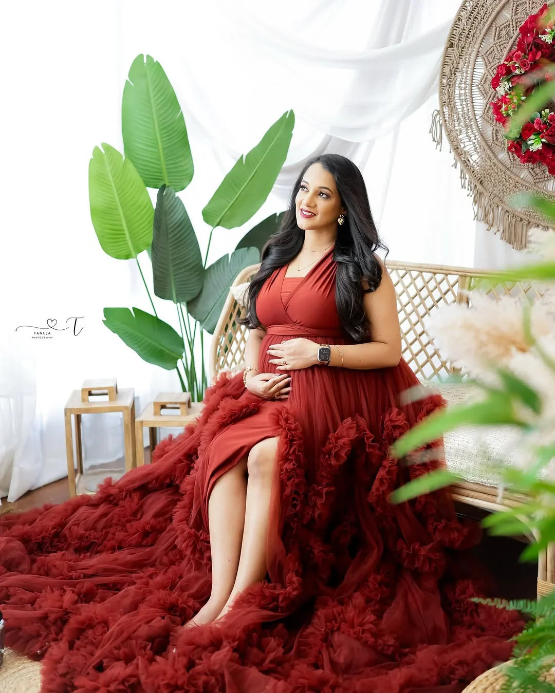Pregnant woman in elegant burgundy maternity gown sitting on rattan bench in bright, leafy setting